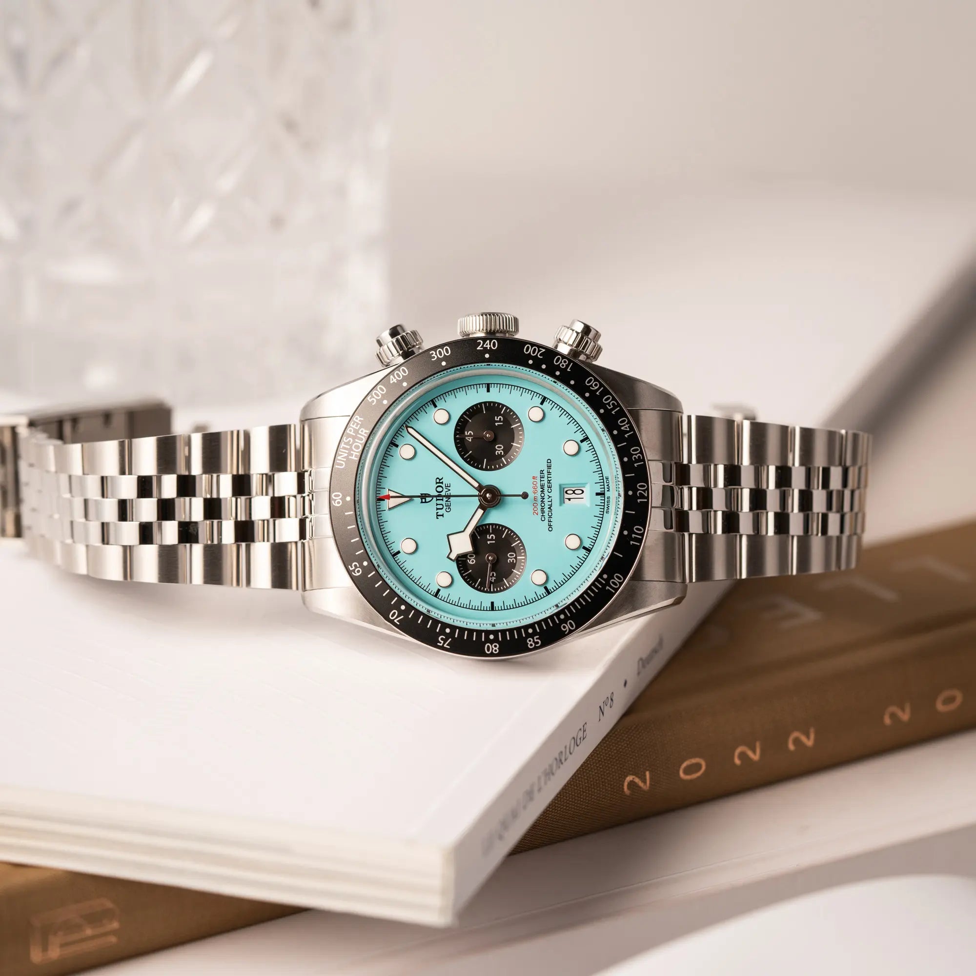 Tudor Black Bay Chronograph Flamingo mit dem türkis-blauen Zifferblatt mit schwarzen Totalisatoren und schwarzer Lünette mit Zähler-Skala liegt seitlich auf einem Buch über Uhren