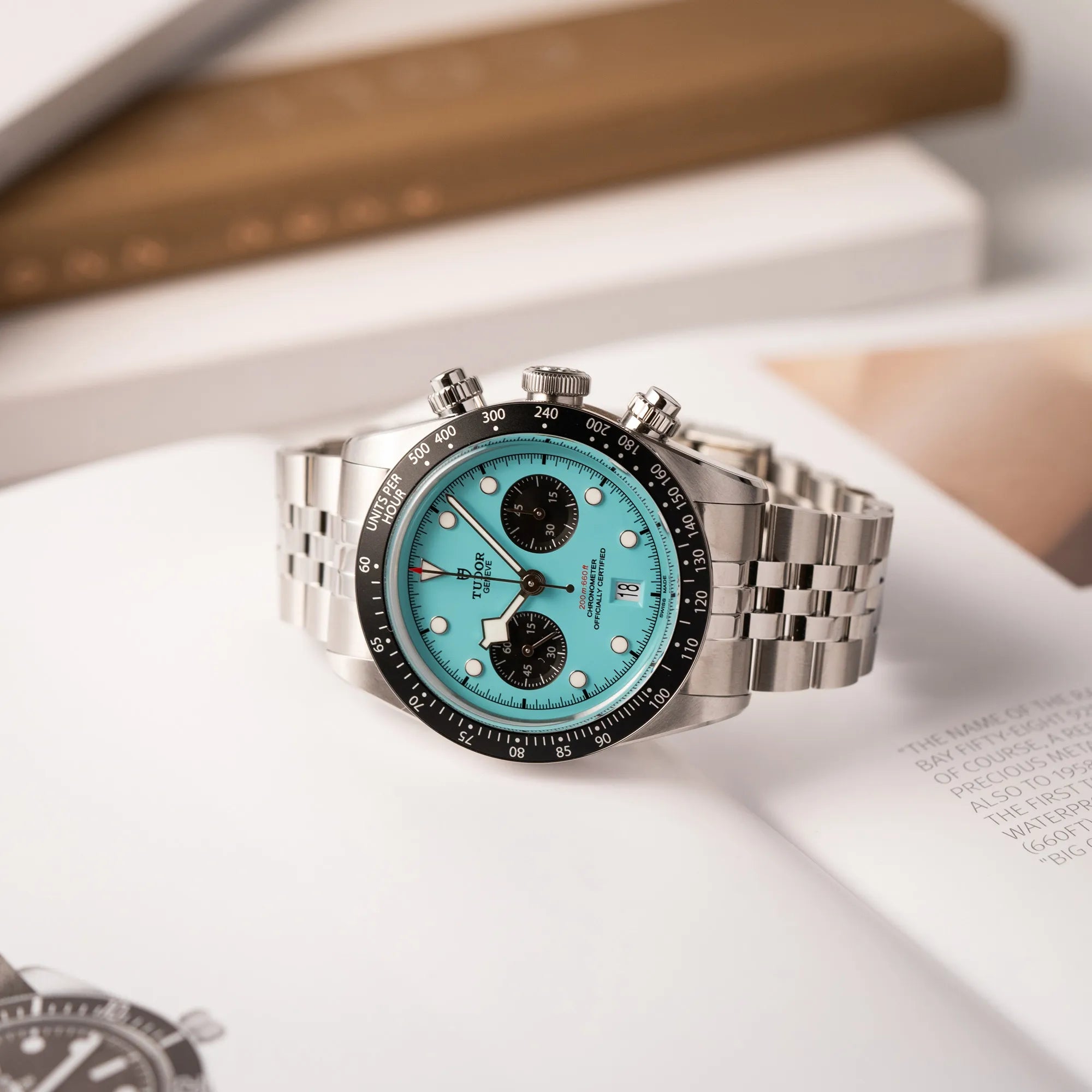 Produktfotografie des Tudor Black Bay Chronographs Flamingo mit dem türkis-blauen Zifferblatt mit schwarzen Totalisatoren und schwarzer Lünette mit Zähler-Skala, während die Uhr auf dem Tudor-Katalog liegt