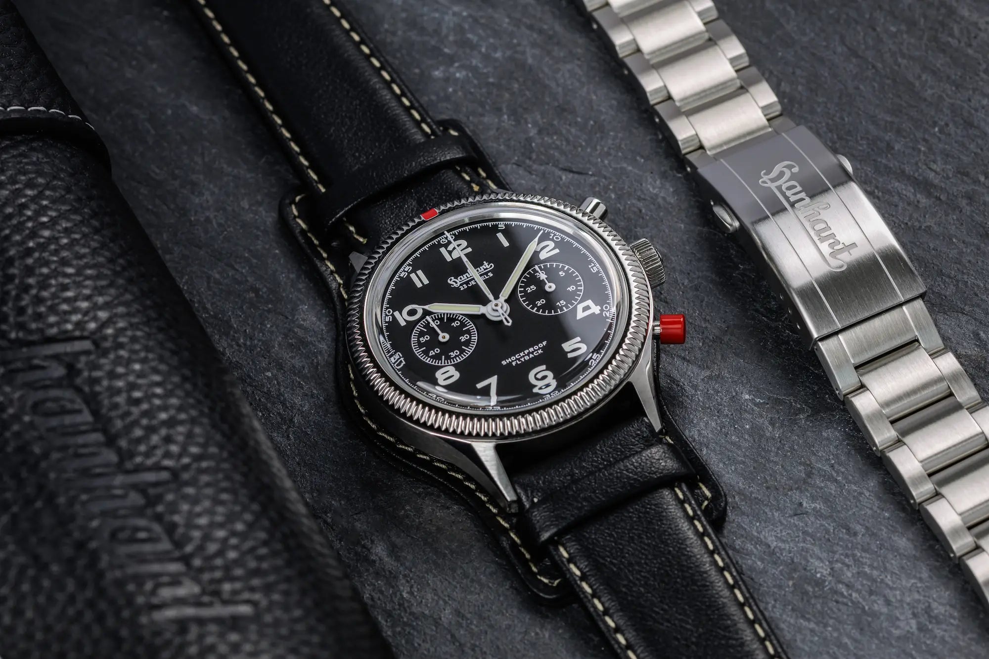 Produktfotografie der Hanhart 417 ES 1954 Heritage Flyback mit 39mm-Gehäuse und schwarzem Zifferblatt am schwarzen Lederband, während das Edelstahlarmband daneben liegt