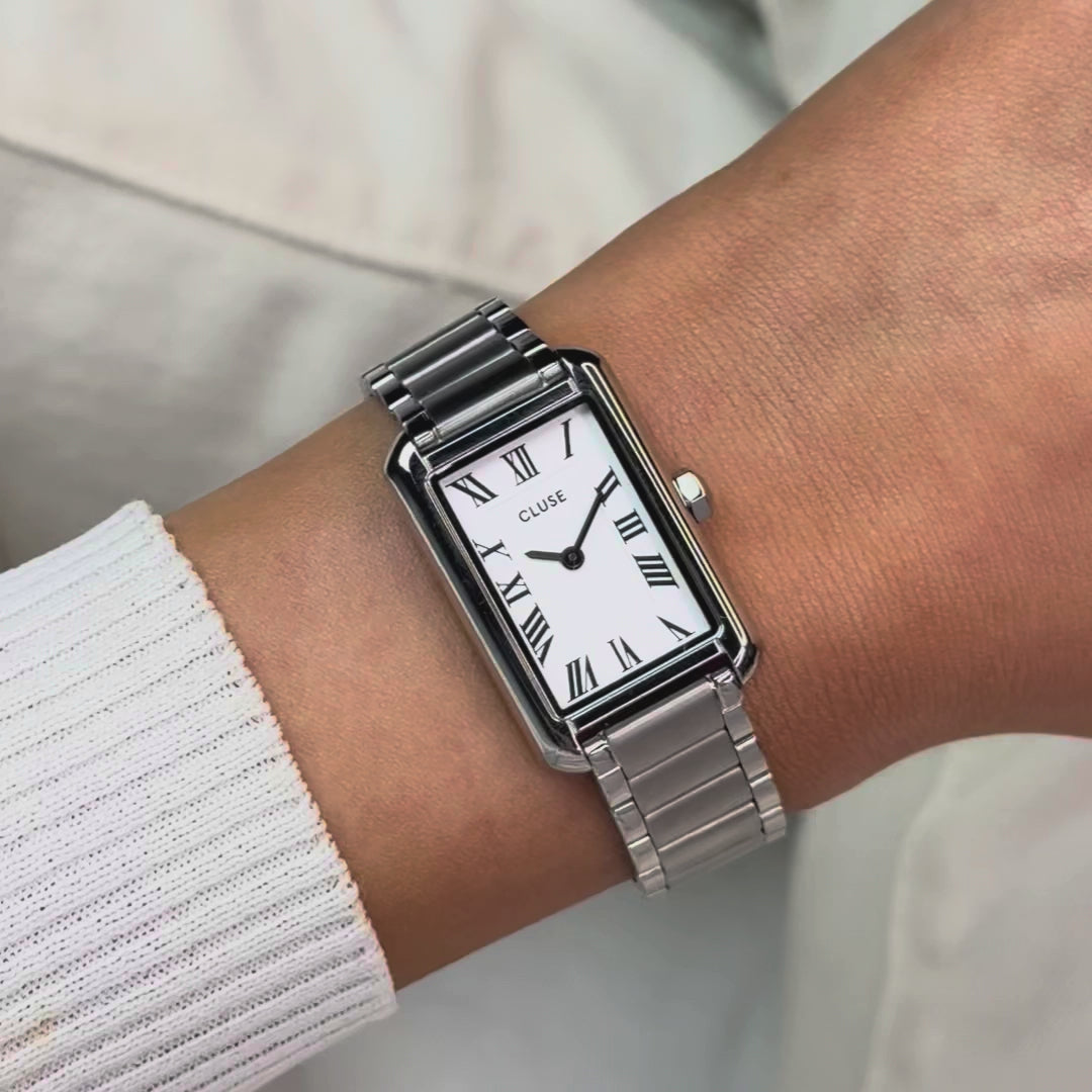 Frau trägt die silberfarbene Cluse Belisenna Damenuhr mit länglichem, rechteckigem Gehäuse und weißem Zifferblatt mit römischen Indizes an ihrem Handgelenk und präsentiert die Uhr in die Kamera