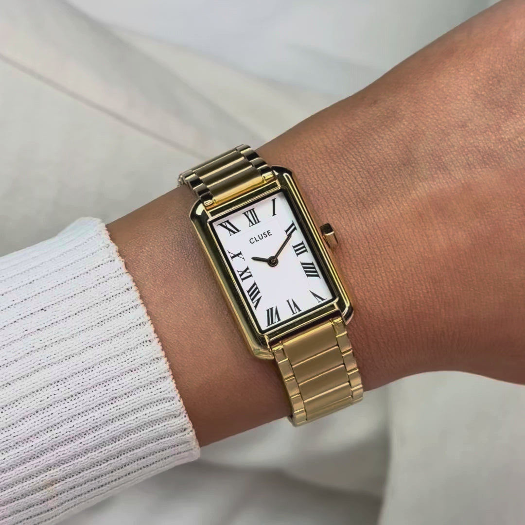 Frau trägt die goldfarbene Cluse Belisenna Damenuhr mit länglichem, rechteckigem Gehäuse und weißem Zifferblatt mit römischen Indizes an ihrem Handgelenk und präsentiert die Uhr in die Kamera