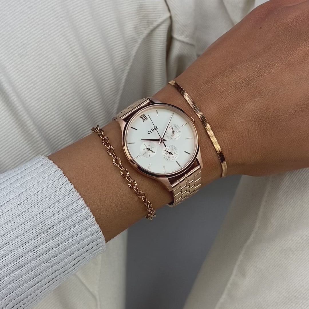 Frau trägt die Cluse Stahl-Uhr "Minuit Multifunction" im Rosègold-Look mit weißem Zifferblatt und drei Sub-Dials zur Wochentags-, Datums-Anzeige und 24 Stunden-Zähler und präsentiert die Uhr in die Kamera