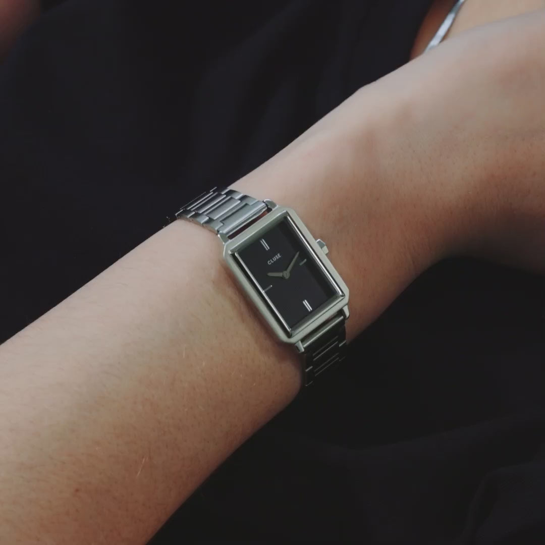 Frau trägt die Cluse "Fluette" Stahl-Uhr mit schwarzem, gestreiften Zifferblatt mit silberfarbenem Gehäuse mit Edelstahl-Armband und kombiniert die Uhr mit silbernen Armbändern und präsentiert diese in die Kamera