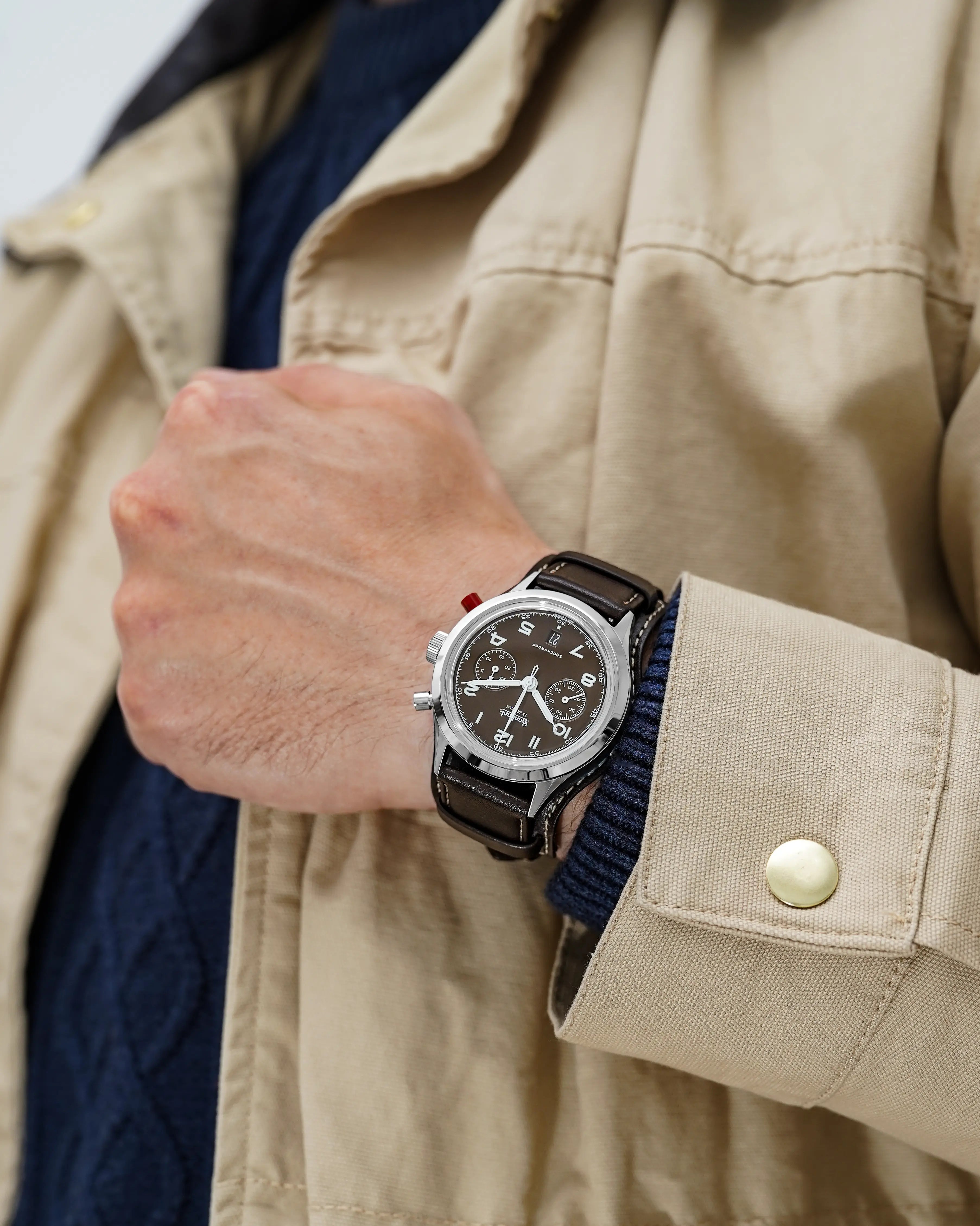 Mann in einer beigen Jacke trägt die Hanhart 417 ES Mokka Limited Edition mit einem braunem Zifferblatt an einem passenden braunem Lederband mit Unterlage an seinem Handgelenk 