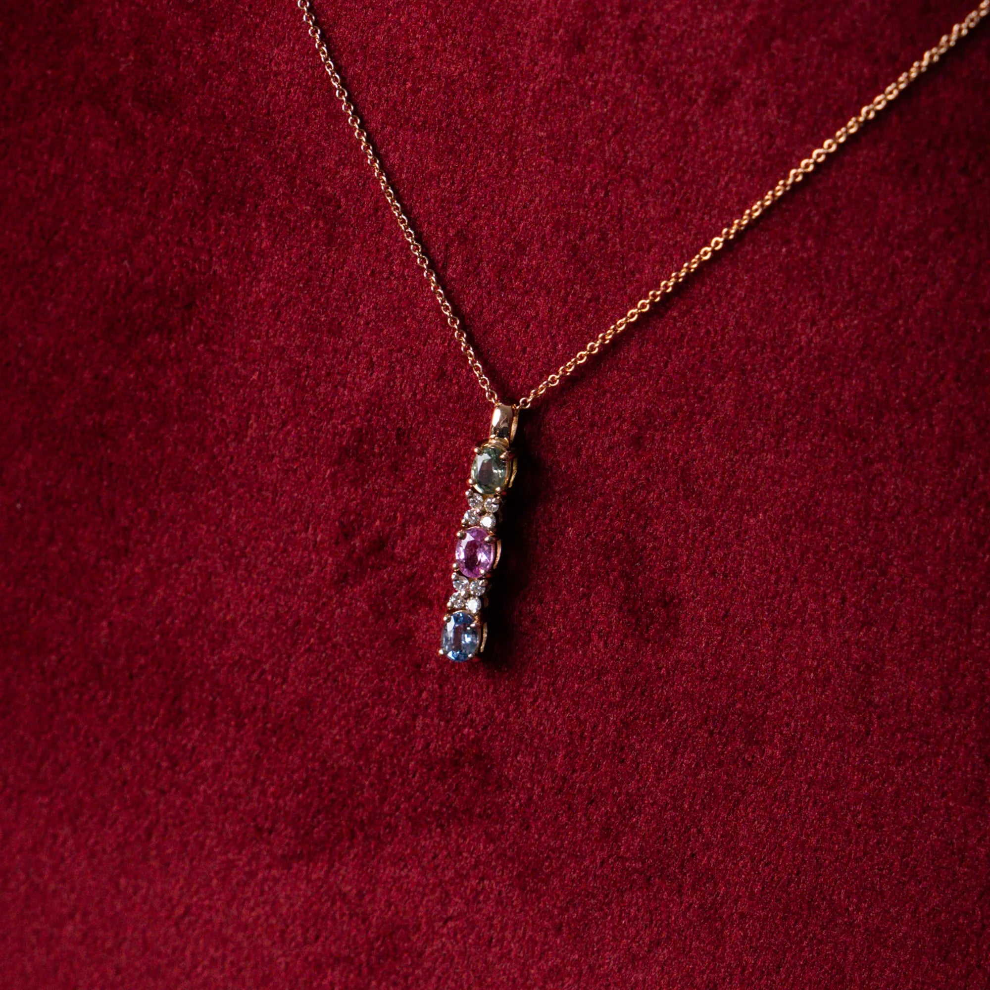 Detailaufnahme des rosegoldenen Colliers CDZM-31R von Lunatica Roma mit drei multicolor Saphiren und dazwischen platzierten Diamant-Blüten, während dieses auf einem samtigen, weinroten Ständer präsentiert wird