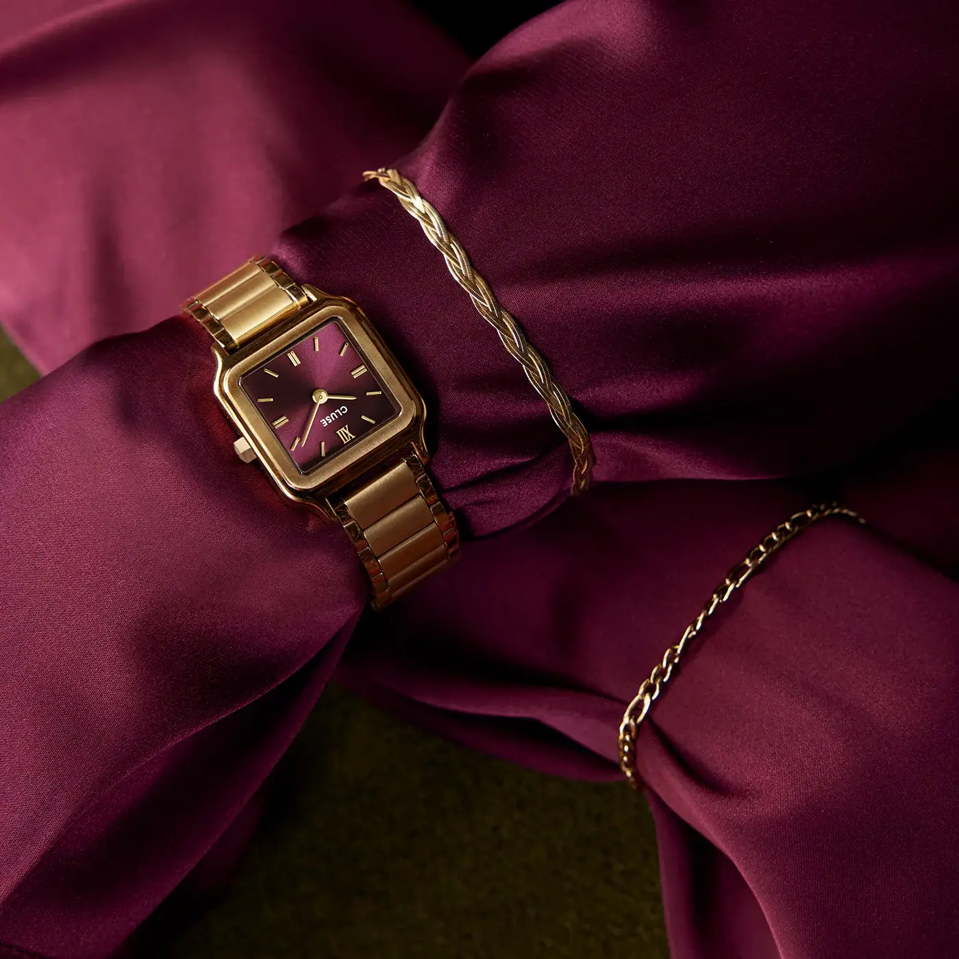Detailaufnahme vom Handgelenk einer Frau in einem weinroten Kleid zeugt die goldfarbene Cluse Gracieuse Petite Damenuhr mit rechteckigem Gehäuse und dunkelrotem Zifferblatt