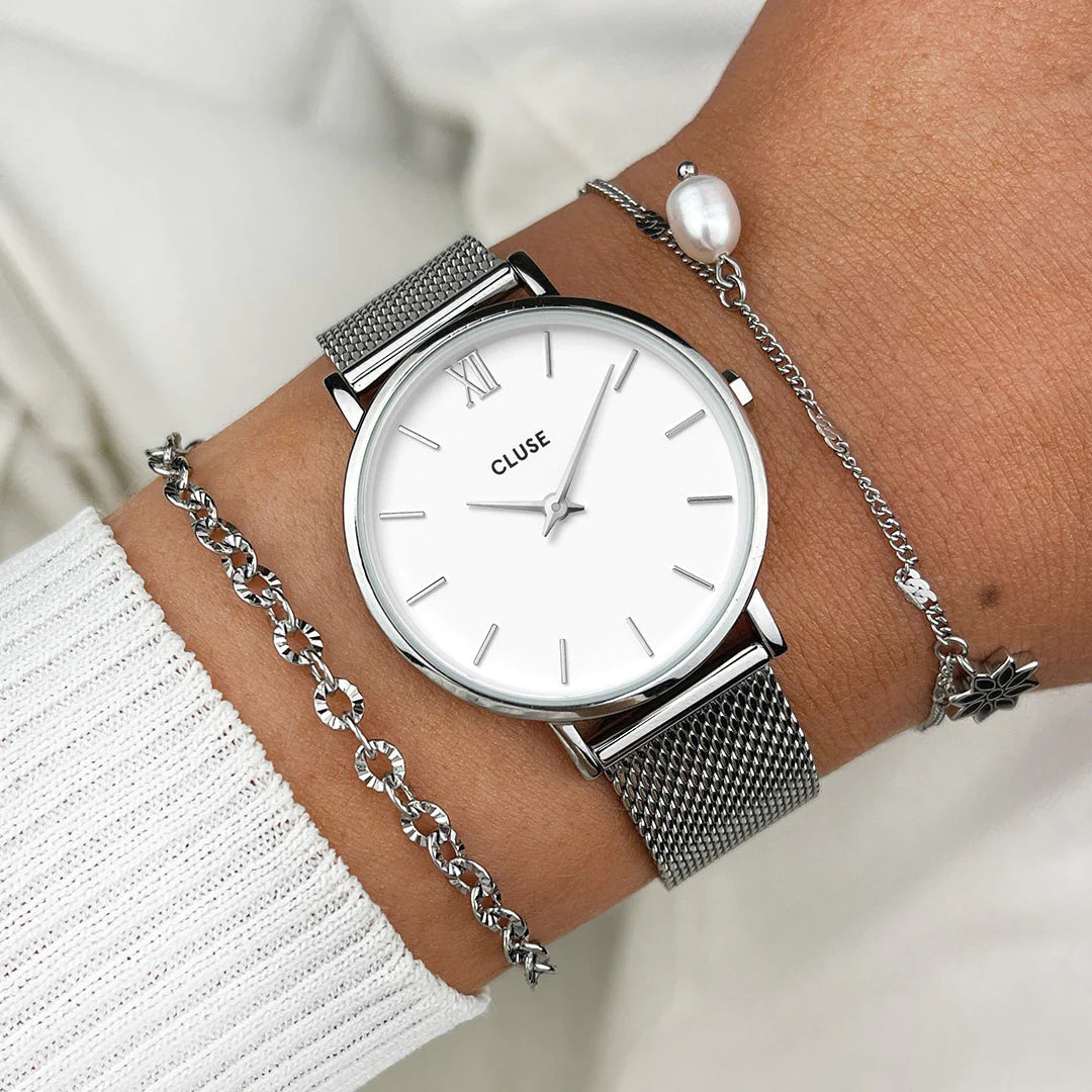 Frau trägt die Cluse Stahl-Uhr "Minuit Mesh" mit silberfarbenem Gehäuse und weißem Zifferblatt an ihrem Handgelenk und kombiniert diese mit zwei silbernen Armbändern