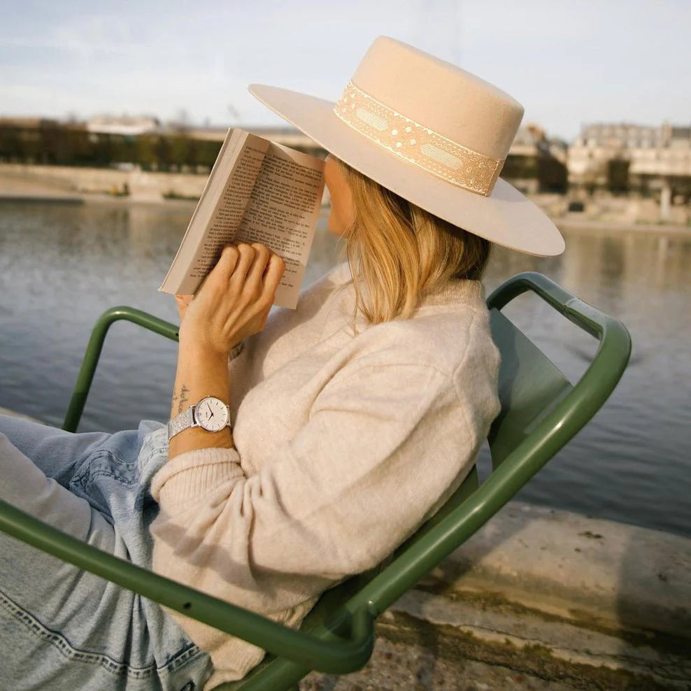 Frau mit Hut sitzt am Wasser und liest in einem grünen Stuhl ein Buch während sie die Cluse Stahl-Uhr "Minuit Mesh" mit silberfarbenem Gehäuse und weißem Zifferblatt an ihrem Handgelenk trägt