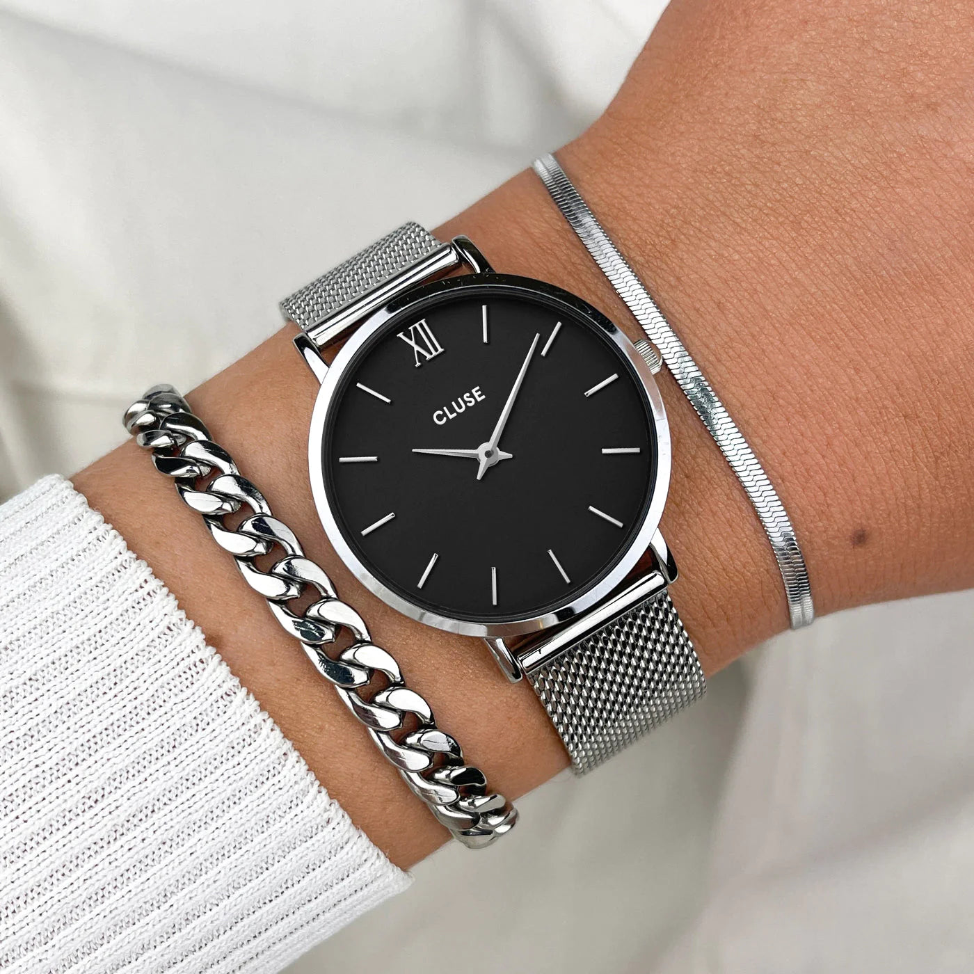 Frau trägt die Cluse Stahl-Uhr "Minuit Mesh" mit silberfarbenem Gehäuse und schwarzem Zifferblatt und kombiniert diese mit zwei silbernen Armbändern