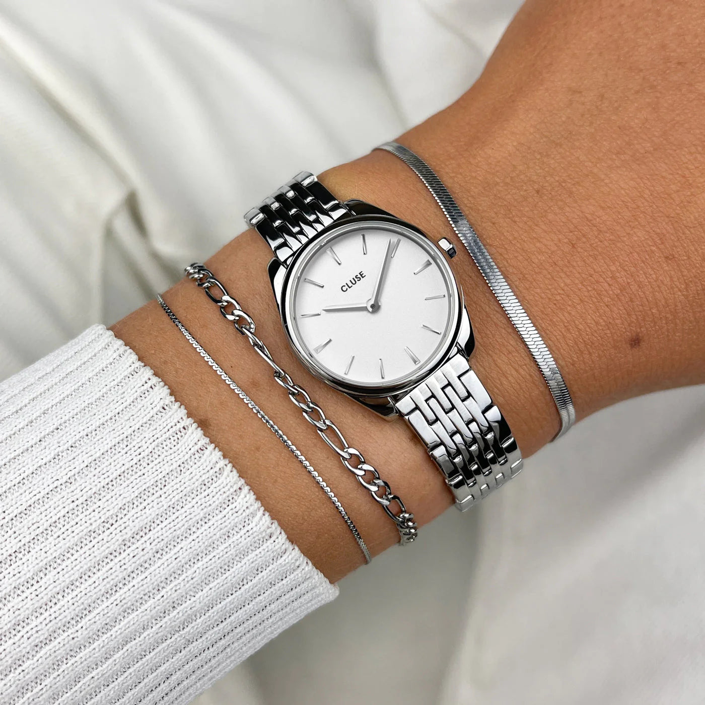 Frau trägt die Cluse Uhr "Feroce Mini" mit silberfarbenem Gehäuse und weißem Zifferblatt am Handgelenk und kombiniert silberne Armbänder damit