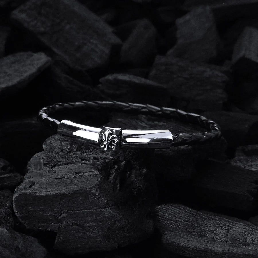 Feines, schwarzes, geflochtenes Lederarmband mit glattem Silberverschluss, welcher durch ein Lilien-Detail  verziert wurde, liegt auf schwarzer Holzkohle