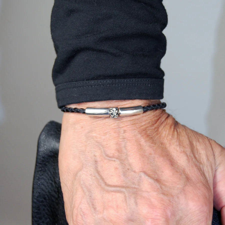 Mann trägt ein feines, schwarzes, geflochtenes Lederarmband mit glattem Silberverschluss, welcher durch ein Lilien-Detail  verziert wurde, am Handgelenk