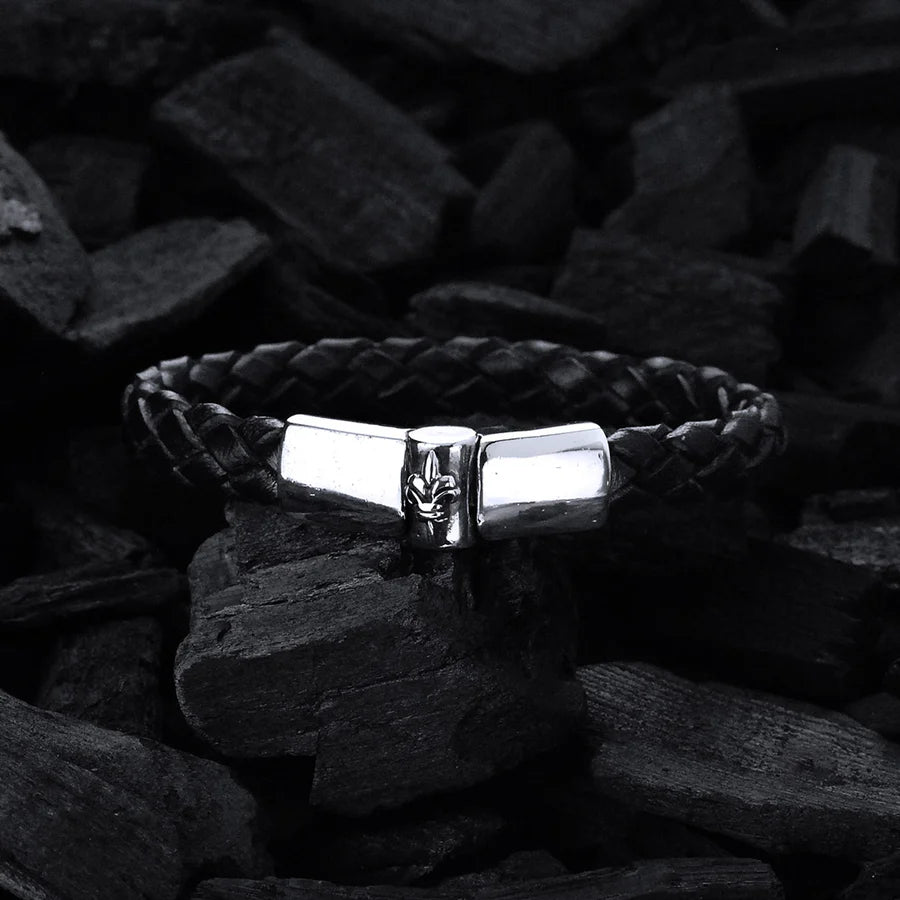 Produktfoto des schwarzen, geflochtenen Lederarmbands mit glattem Silberverschluss  und einer zentralen Lilie als Design-Highlight des Verschlusses vor schwarzer Holzkohle