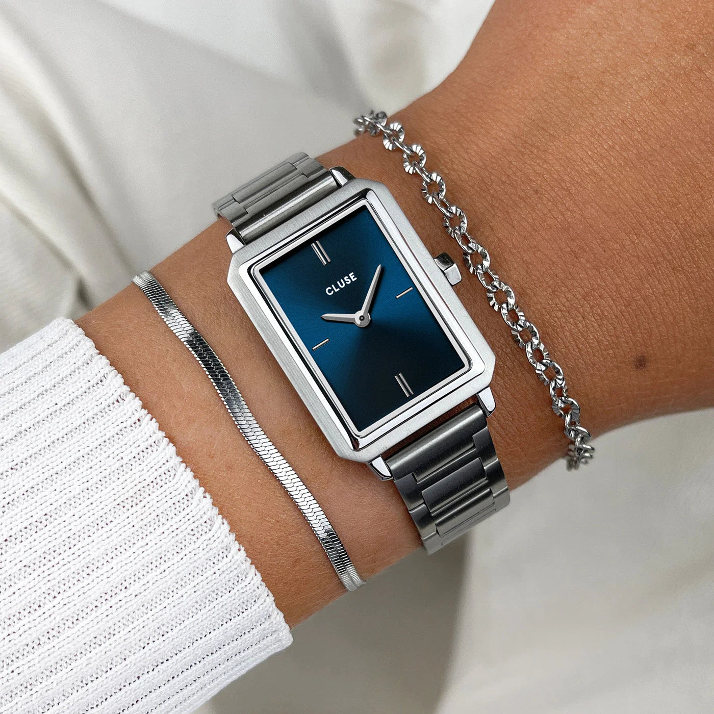 Frau trägt die Cluse "Fluette" Stahl-Uhr mit dunkelblauem Zifferblatt mit silberfarbenem Gehäuse mit Edelstahl-Armband und kombiniert die Uhr mit silbernen Armbändern