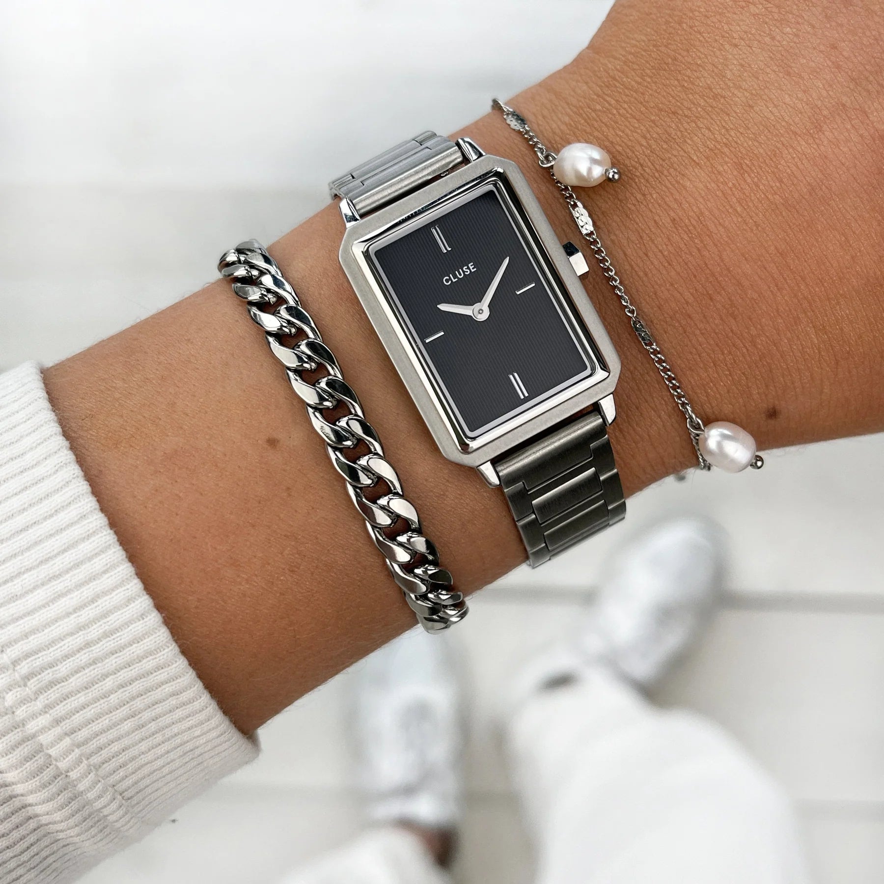 Frau trägt die Cluse "Fluette" Stahl-Uhr mit schwarzem, gestreiften Zifferblatt mit silberfarbenem Gehäuse mit Edelstahl-Armband und kombiniert die Uhr mit silbernen Armbändern
