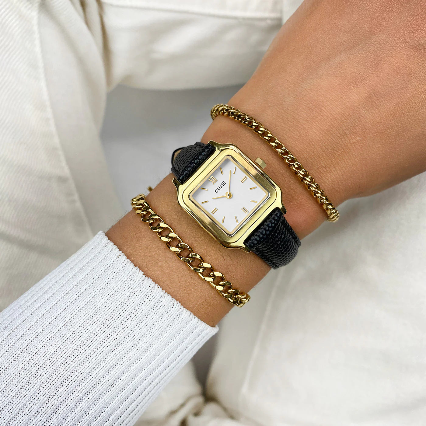 Frau trägt die Cluse Stahl-Uhr "Gracieuse Petite" mit einem gelbgold-farbenem Gehäuse und weißem Zifferblatt am schwarzen Lederband an ihrem Handgelenk