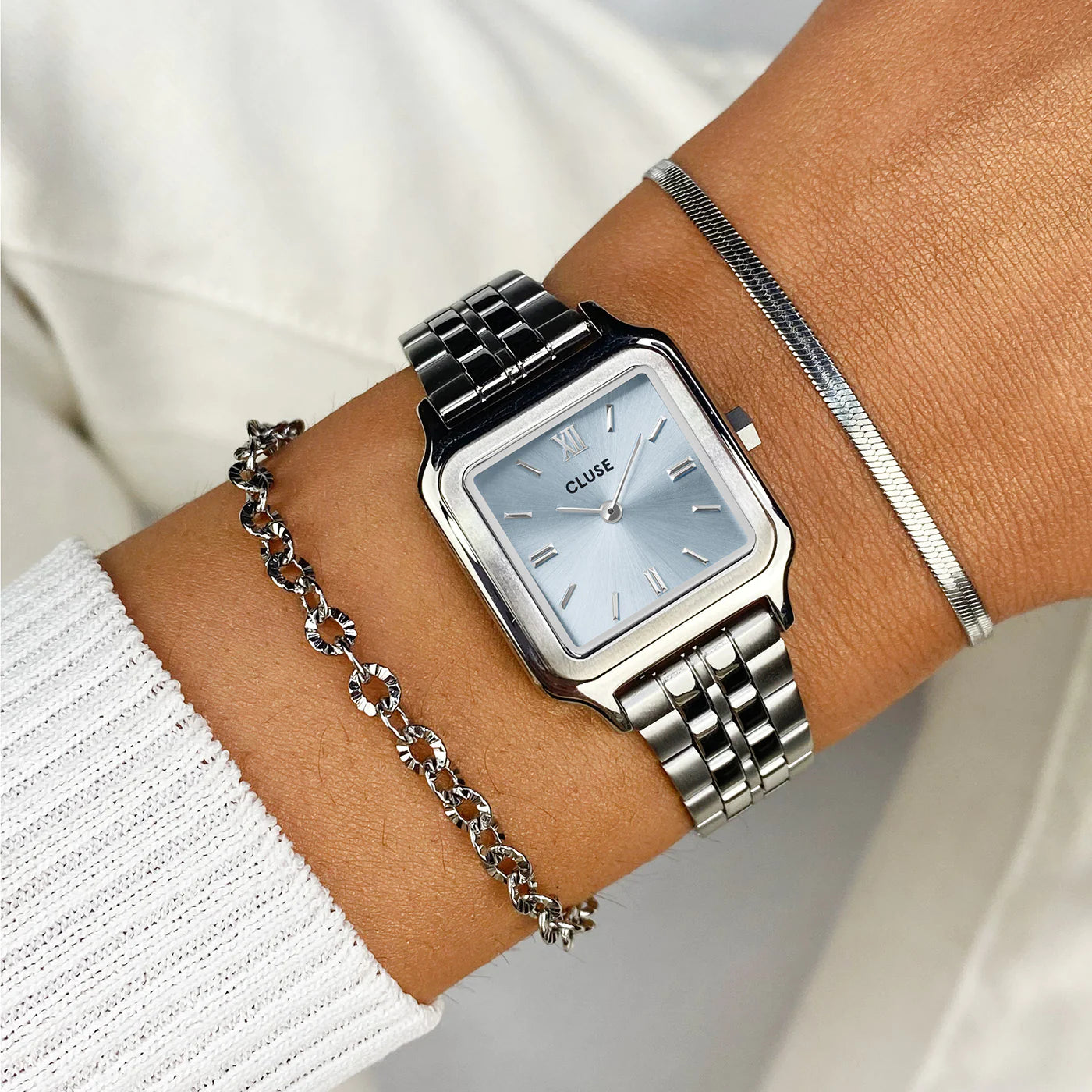 Frau trägt die Cluse Stahl-Uhr "Gracieuse Petite" mit silberfarbenem Gehäuse und eisblauem Zifferblatt an ihrem Handgelenk und kombiniert diese mit zwei silbernen Armbändern