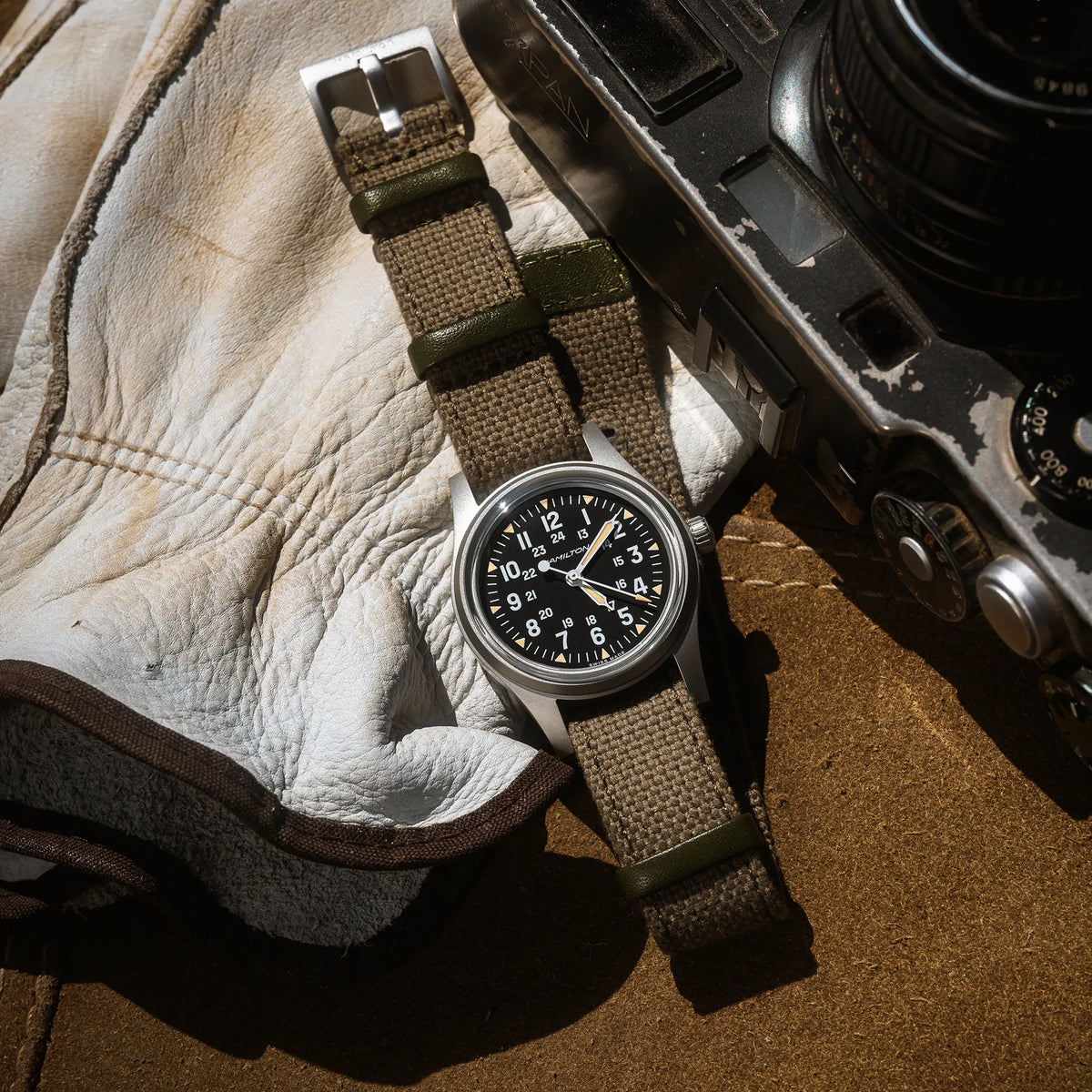Lifestylebild der Hamilton Khaki Field Mechanical H69399930 mit schwarzem Zifferblatt im Vintage-Charakter mit Indizes in weiß und beige an einem khaki-farbenem Nato-Band, neben einem Lederhandschuh und einer Kamera