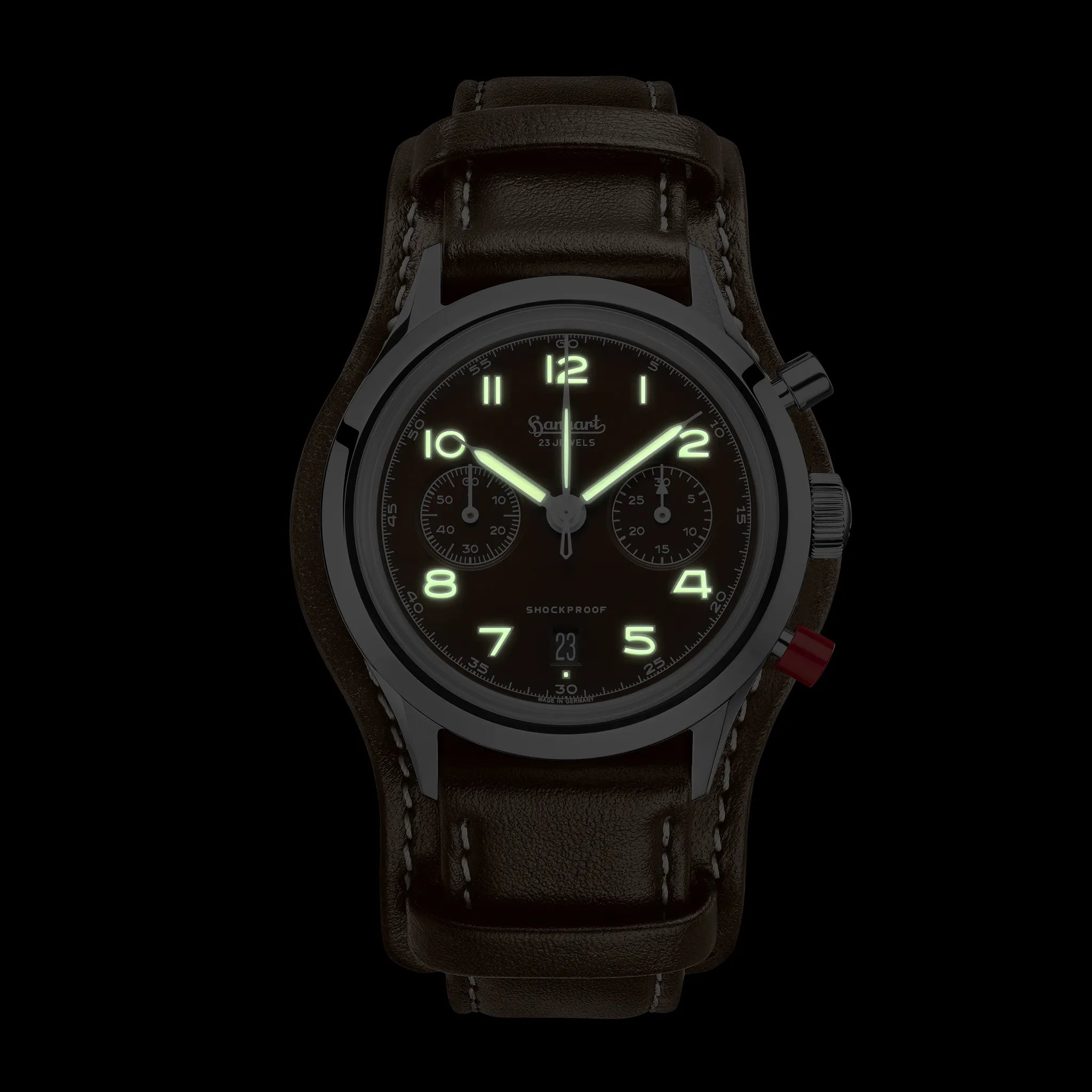 Frontale Ansicht der Hanhart 417 ES Mokka Limited Edition mit einem braunem Zifferblatt an einem passenden braunem Lederband mit Unterlage bei Nacht mit leuchtenden Zeigern und Indizes