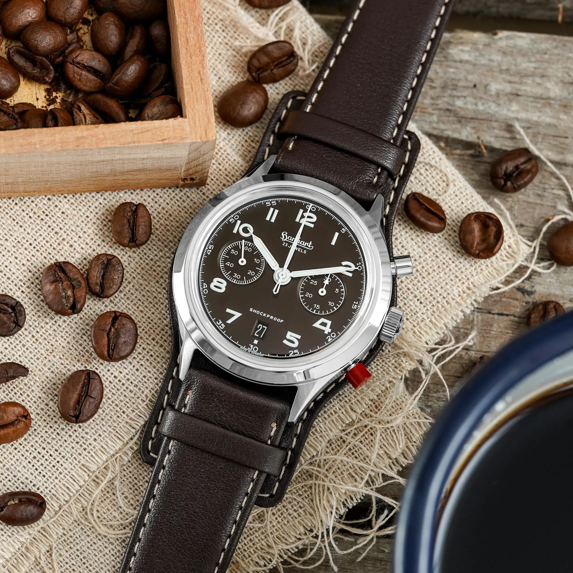 Produktfotografie der Hanhart 417 ES Mokka Limited Edition mit einem braunem Zifferblatt an einem passenden braunem Lederband mit Unterlage neben ebenso braunen Kaffeebohnen