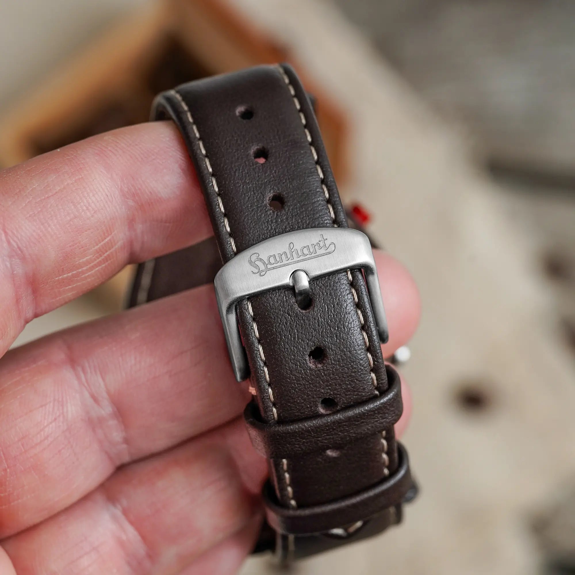 Mann hält die Hanhart 417 ES Mokka Limited Edition mit einem braunem Zifferblatt an einem passenden braunem Lederband mit Unterlage und präsentiert die Dornschliesse in die Kamera