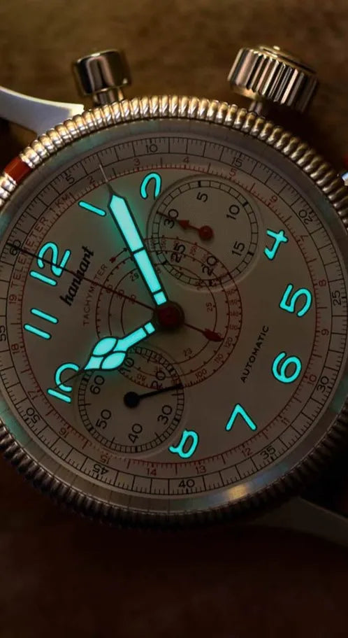 Hanhart Pioneer TachyTele Chronograph mit altweißem Zifferblatt und rot gehaltener Tachymeter-Skala und Telemeter-Skala am braunen Kalbslederband mit Niete bei Dunkelheit zeigt die Funktionalität der hellblau strahlenden Leuchtmasse
