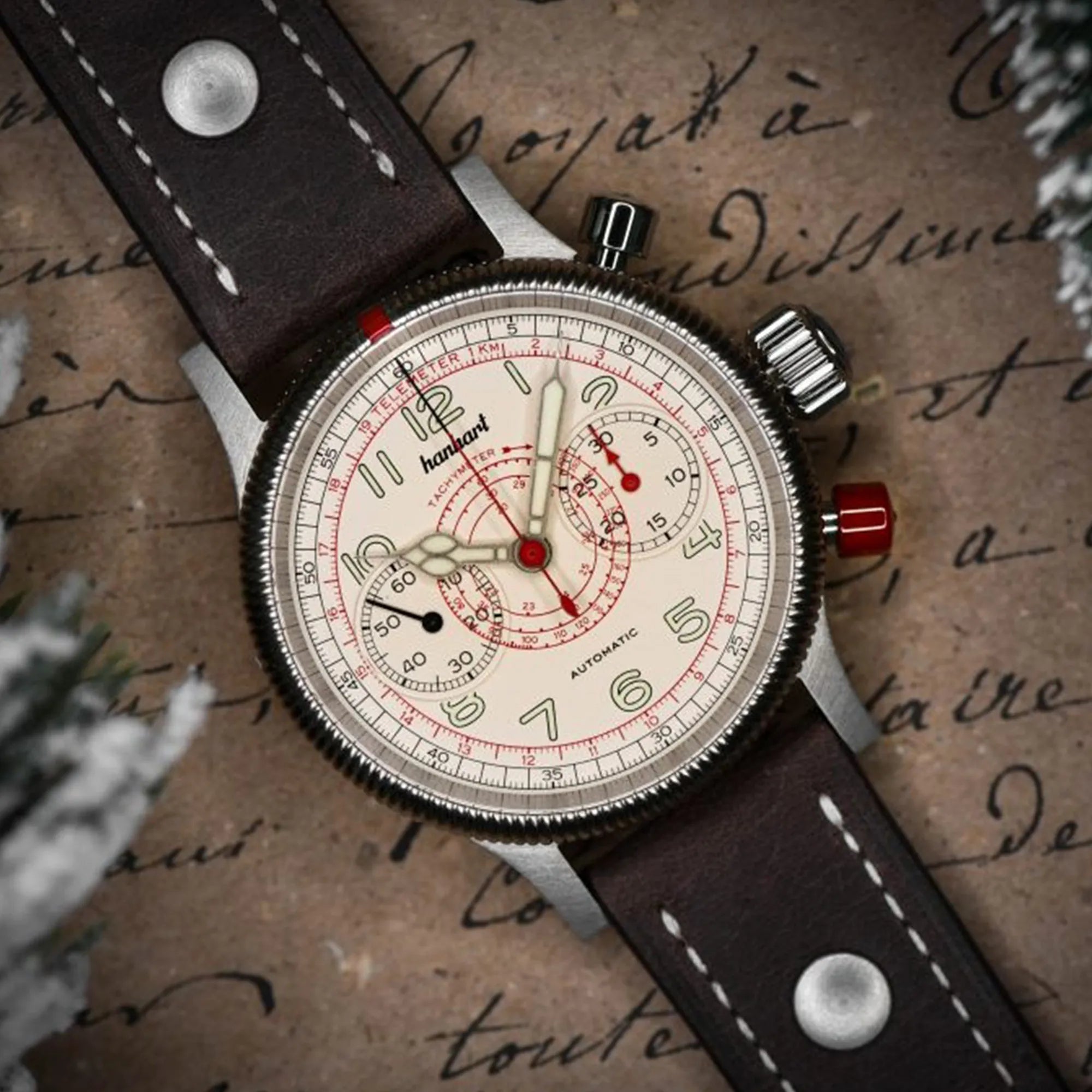 Produktfotografie des Hanhart Pioneer TachyTele Chronograph mit altweißem Zifferblatt und rot gehaltener Tachymeter-Skala und Telemeter-Skala am braunen Kalbslederband mit Niete