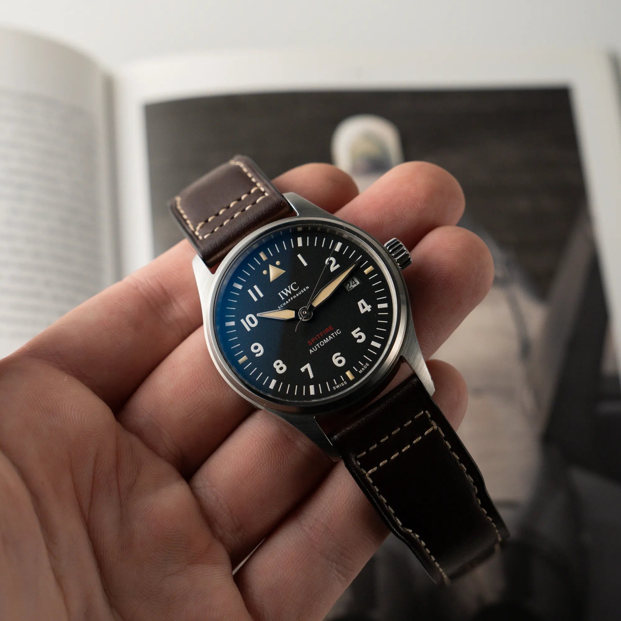 Mann hält die IWC Pilotenuhr Automatic Spitfire IW326803 mit schwarzen Zifferblatt an einem braunem Lederband in seiner Hand und zeigt die Armbanduhr in die Kamera