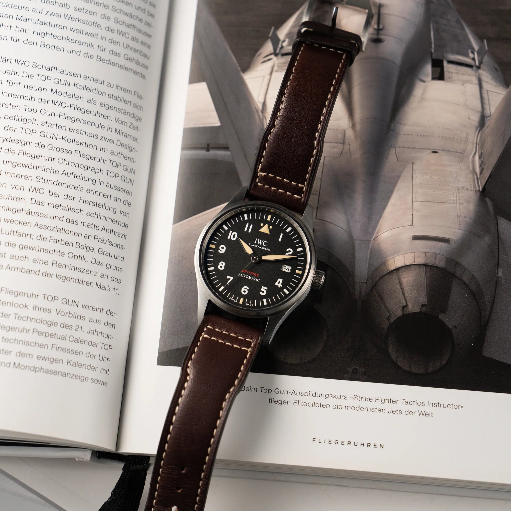 Produktfotografie der IWC Pilotenuhr Automatic Spitfire IW326803 mit schwarzen Zifferblatt an einem braunem Lederband, während die Uhr auf einem Buch über Fliegeruhren liegt