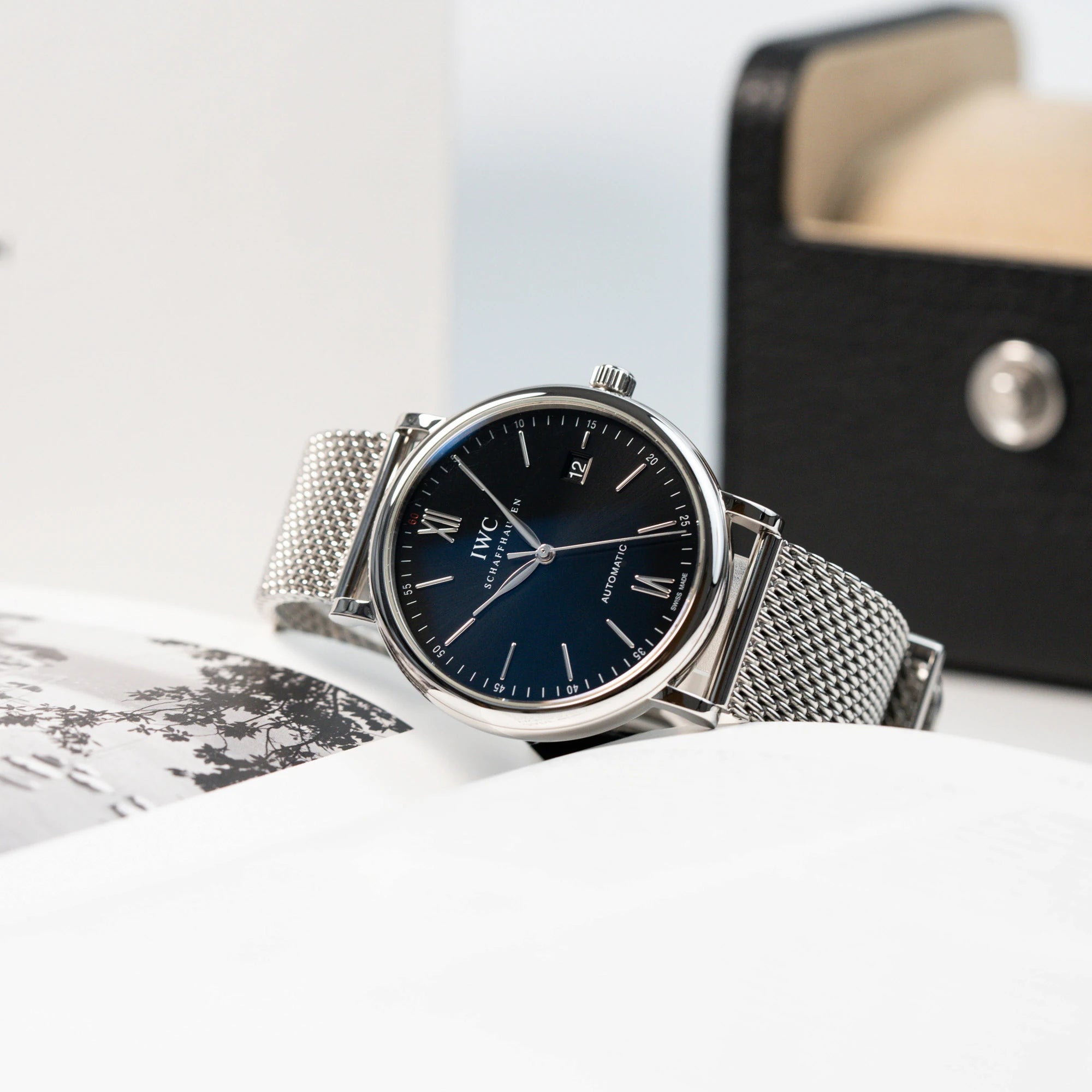 Produktfotografie der IWC Portofino 40 Automatik in Edelstahl mit schwarzem Zifferblatt, während die Uhr an einem aufgeschlagenen Buch lehnt