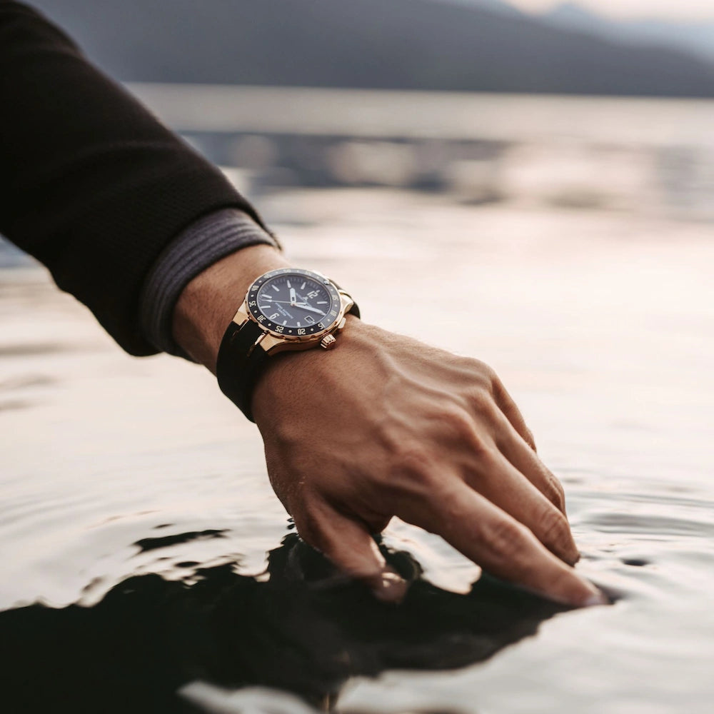 Mann streckt seine Hand ins Wasser und trägt dabei eine Jacques Lemans Uhr mit rosegold-farbenem Gehäuse am Handgelenk