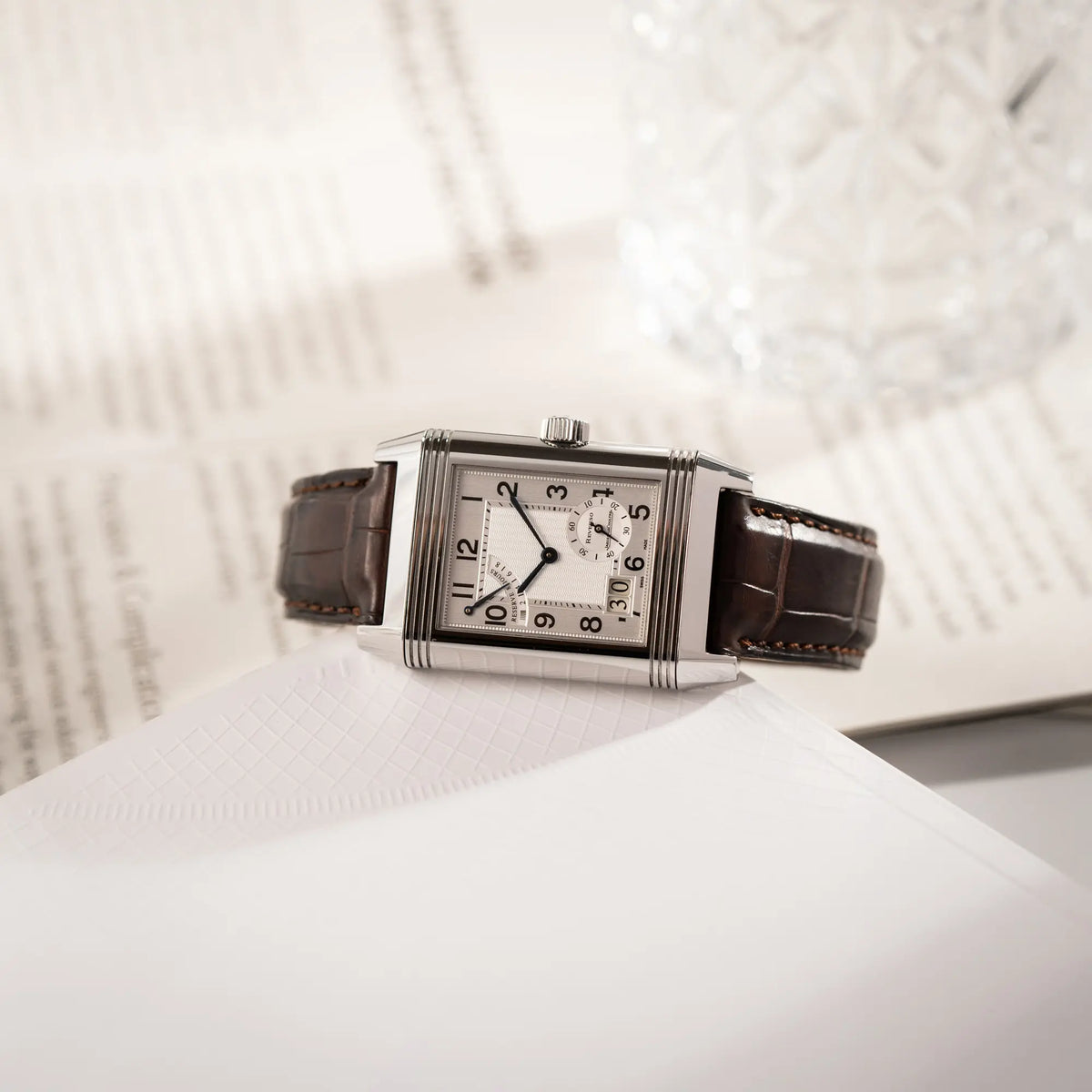Produktfotografie der Jaeger-LeCoultre Reverso Grande Date der Referenz 240.8.15 mit silberfarbenem Zifferblatt und Großdatums-Anzeige, kleiner Sekunde und Gangreserve-Anzeige am braunen Lederband, während die Uhr an einem Buch liegt