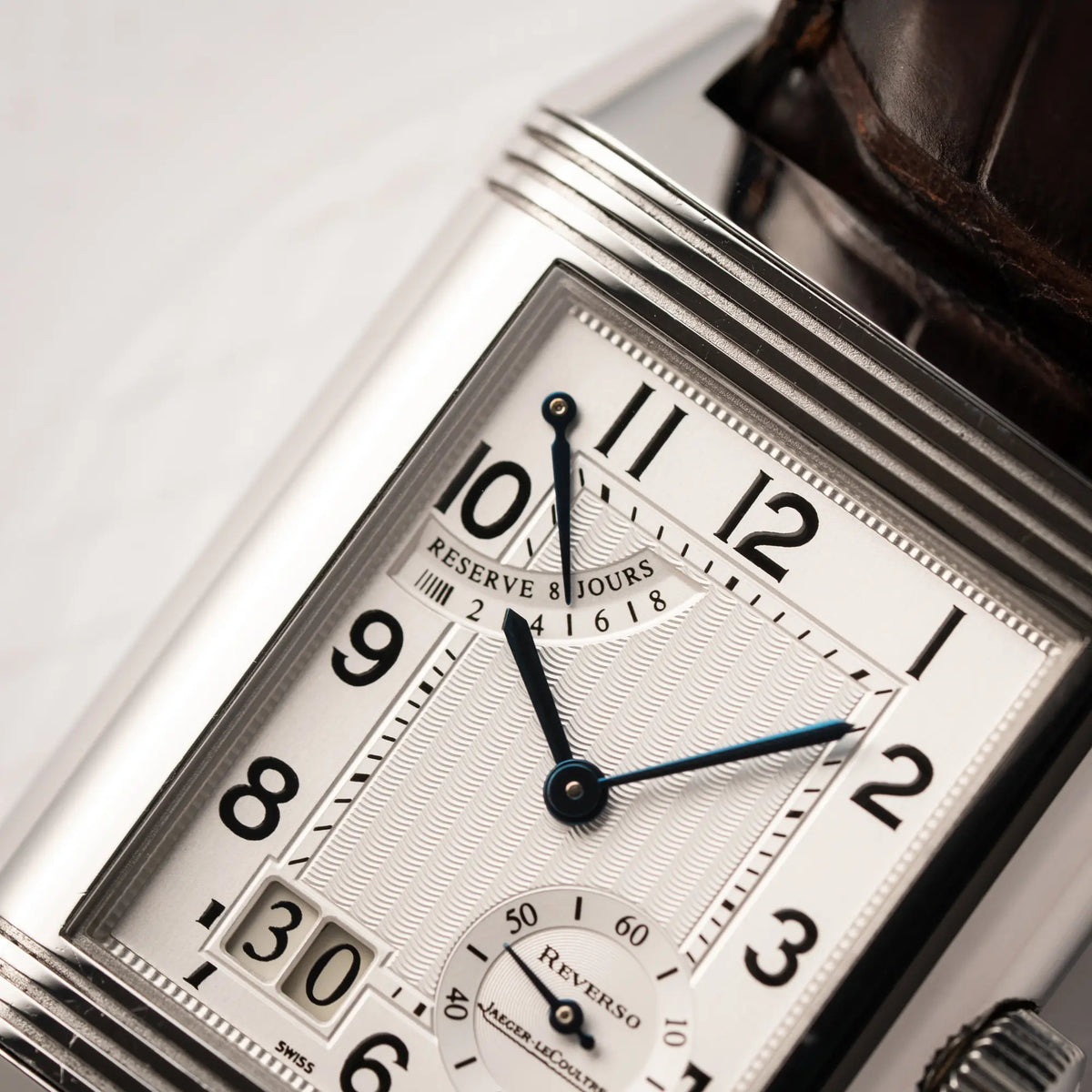 Nahaufnahme der Jaeger-LeCoultre Reverso Grande Date der Referenz 240.8.15 mit silberfarbenem Zifferblatt und Großdatums-Anzeige, kleiner Sekunde und Gangreserve-Anzeige am braunen Lederband, zeigt das Guilloche-Muster des Zifferblatts und die gebläuten Zeiger