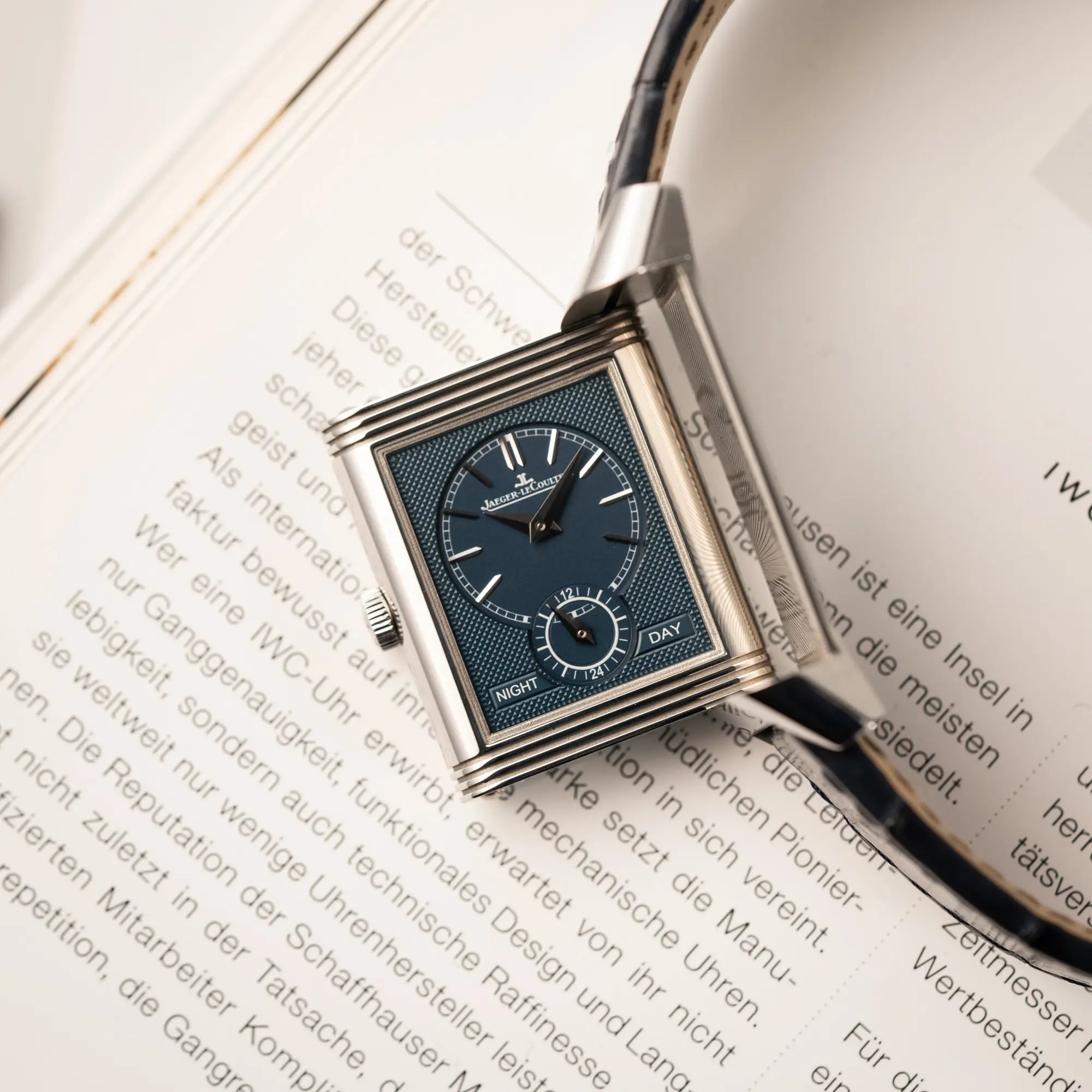 Detailaufnahme der Jaeger-LeCoultre Reverso Tribute Duoface Q3908420 an einem dunkelblauen Lederband zeigt den Reverso-Mechanismus in Bewegung un das blaue Day&Nght-Zifferblatt
