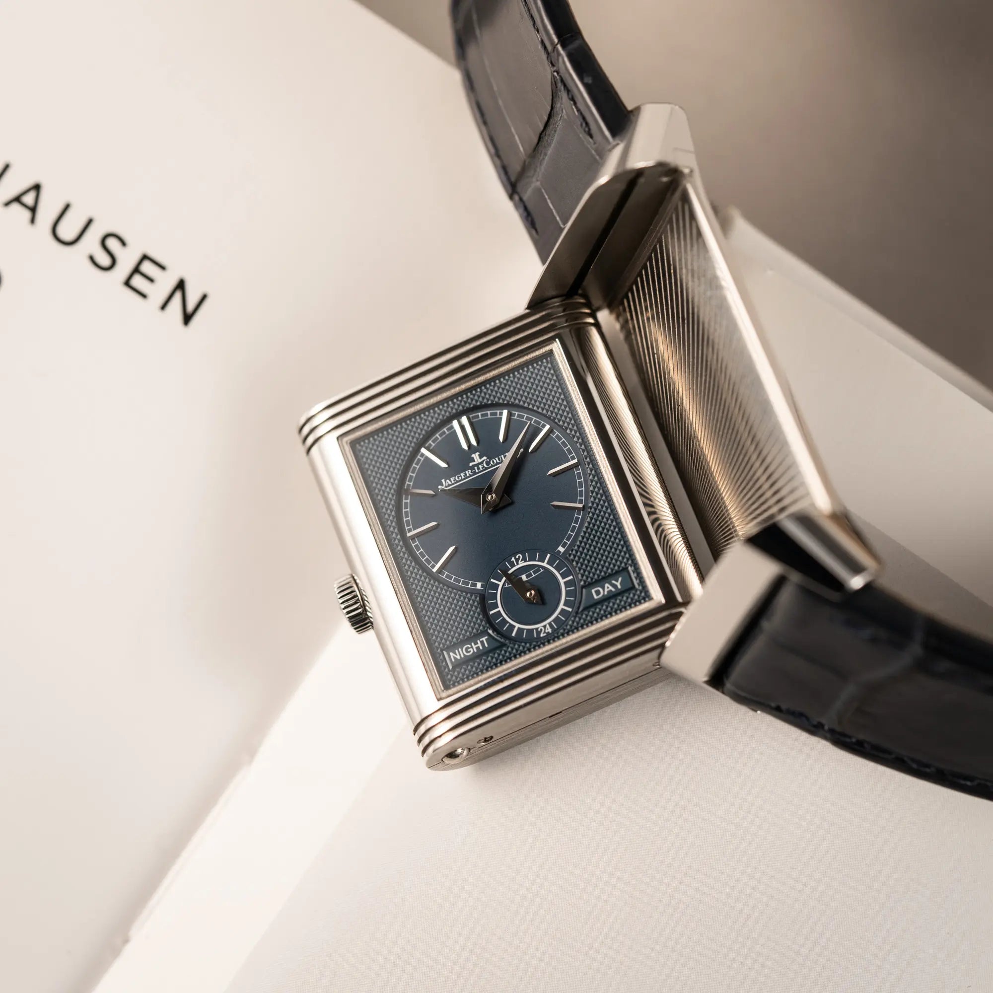 Produktfotografie der Jaeger-LeCoultre Reverso Tribute Duoface Q3908420 an einem dunkelblauen Lederband zeigt den Reverso-Mechanismus in Bewegung un das blaue Day&Nght-Zifferblatt