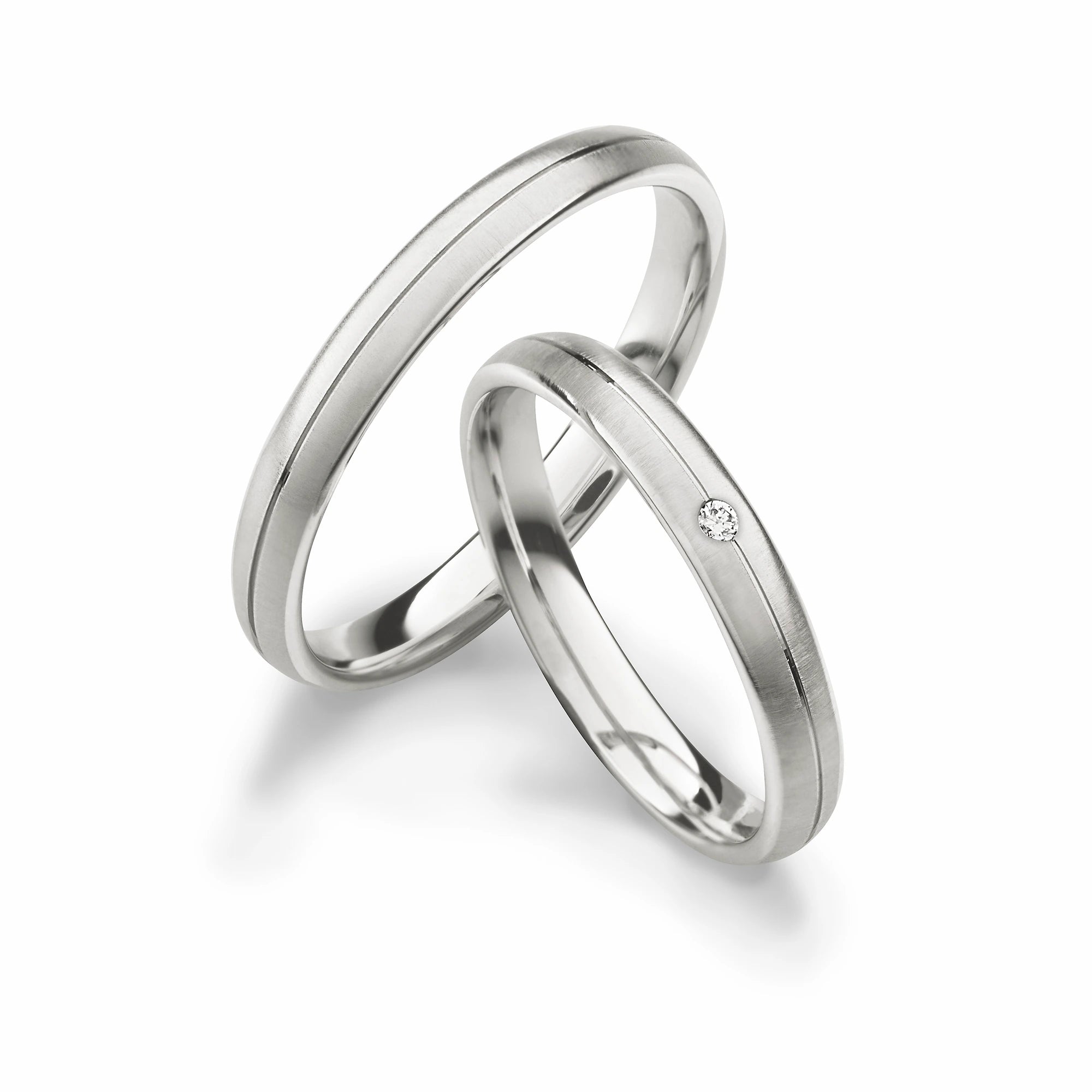 Produktfotografie des weissgoldenen Max Kemper Trauringpaars mit quer gebürsteter Oberflächenstruktur und einer zentralen eingefrästen Nut, sowie einem eingefasstem Brillant als Verzierung des Damenrings