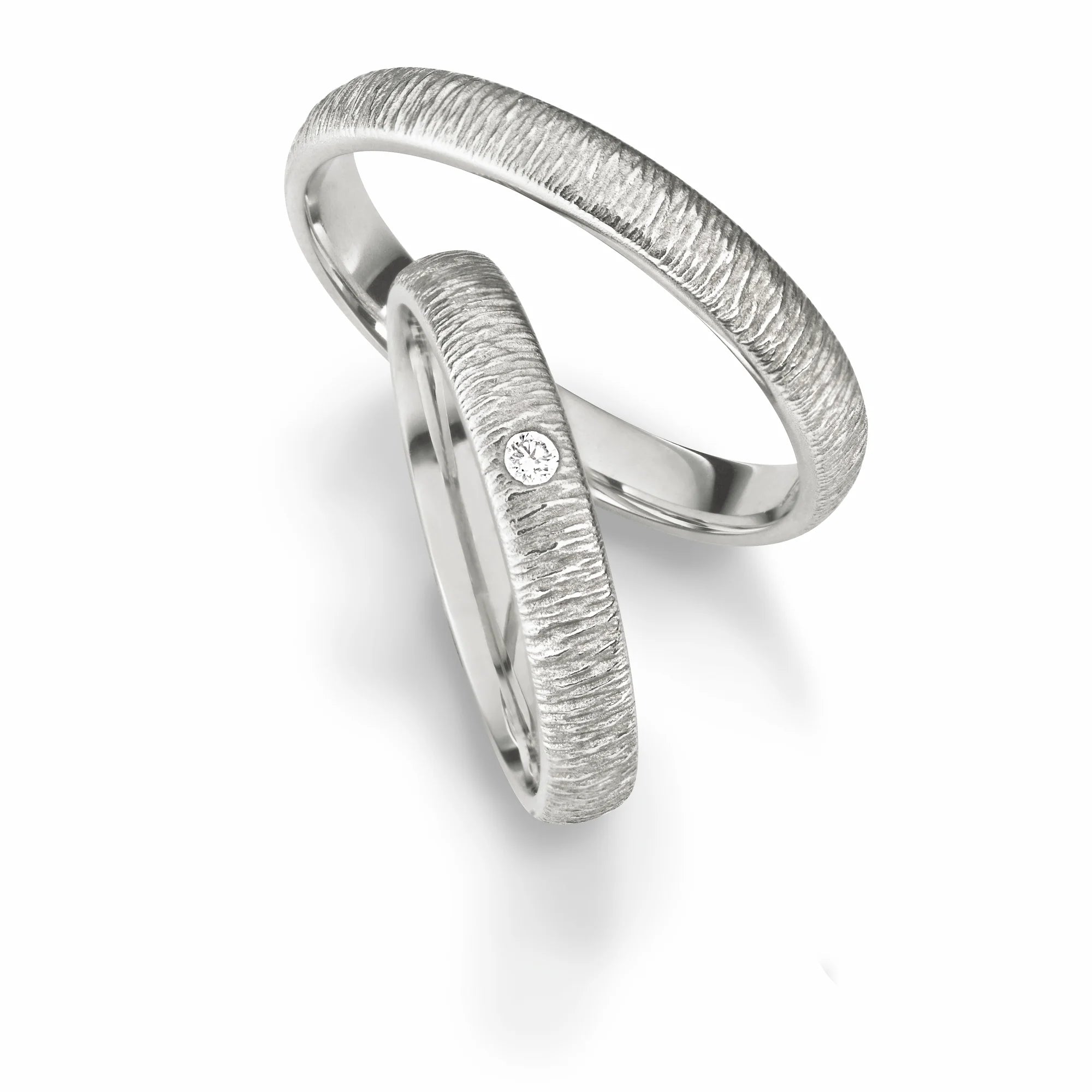 Produktfotografie des weissgoldenen Max Kemper Trauringpaars mit quer verlaufenden Borke-Oberflächenstruktur und einem zentralen Diamanten als Verzierung des Damenrings