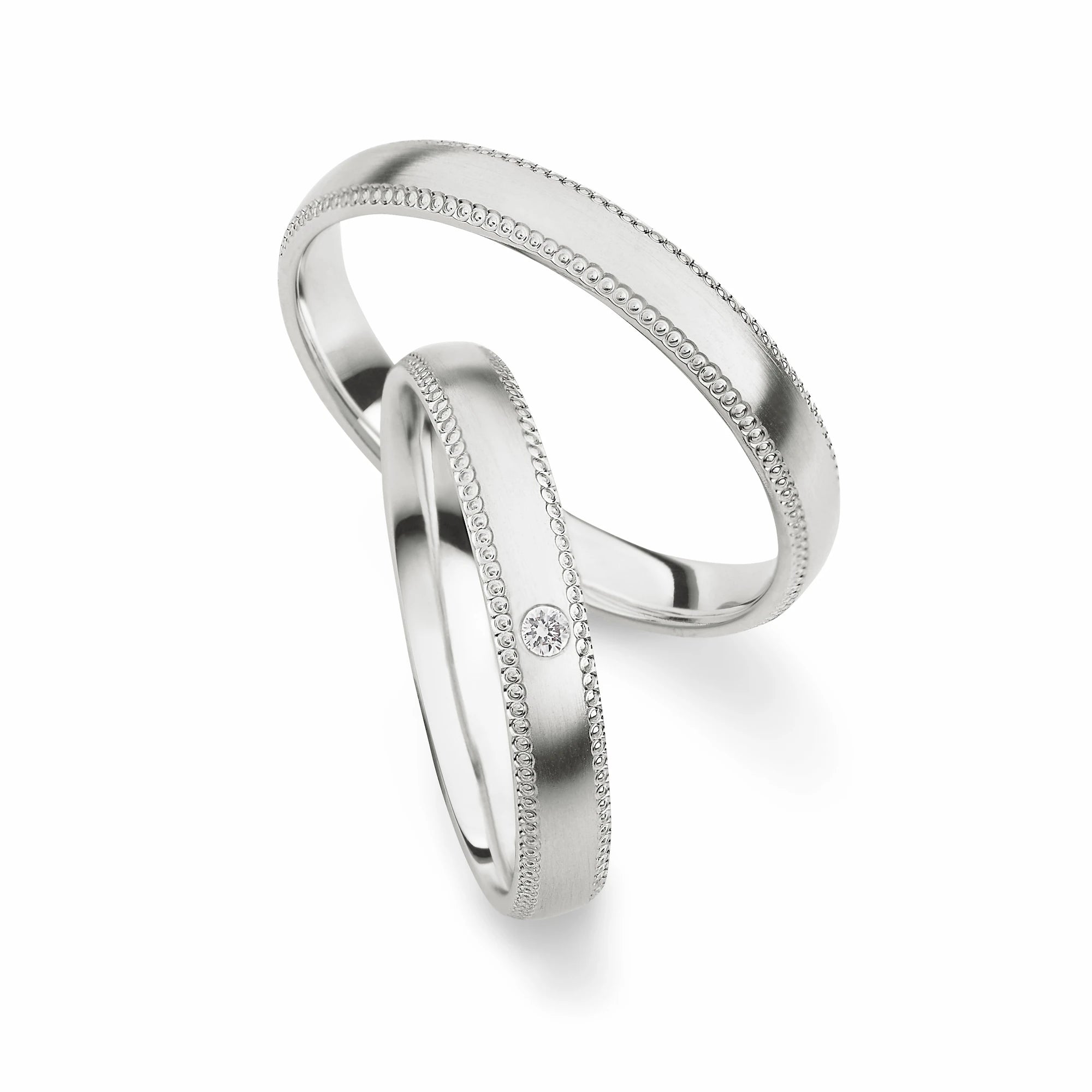 Produktfotografie des weißgoldenen Max Kemper Trauringpaars mit polierter Oberfläche und ornamentartiger Verzierung an den Aussenseiten des Rings , sowie einem Brillanten als Verzierung des Damenrings