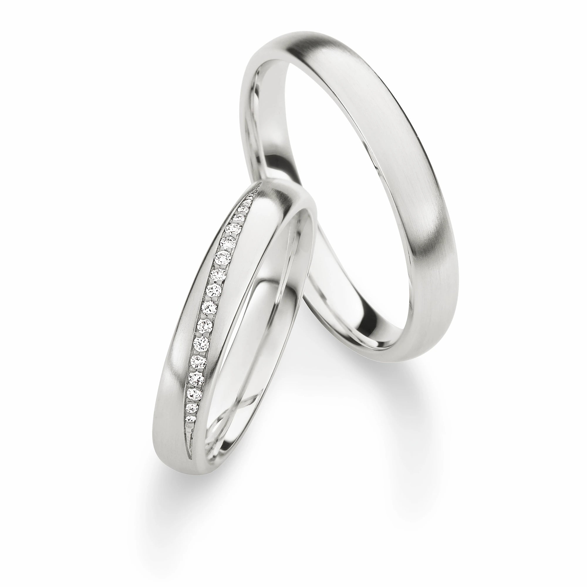 Produktfotografie des weißgoldenen Max Kemper Trauringpaars mit einer glatten polierten Oberflächenstruktur und speziell für den Damenring eingefassten Diamant-Reihe die schräg auf der Ringschiene liegt