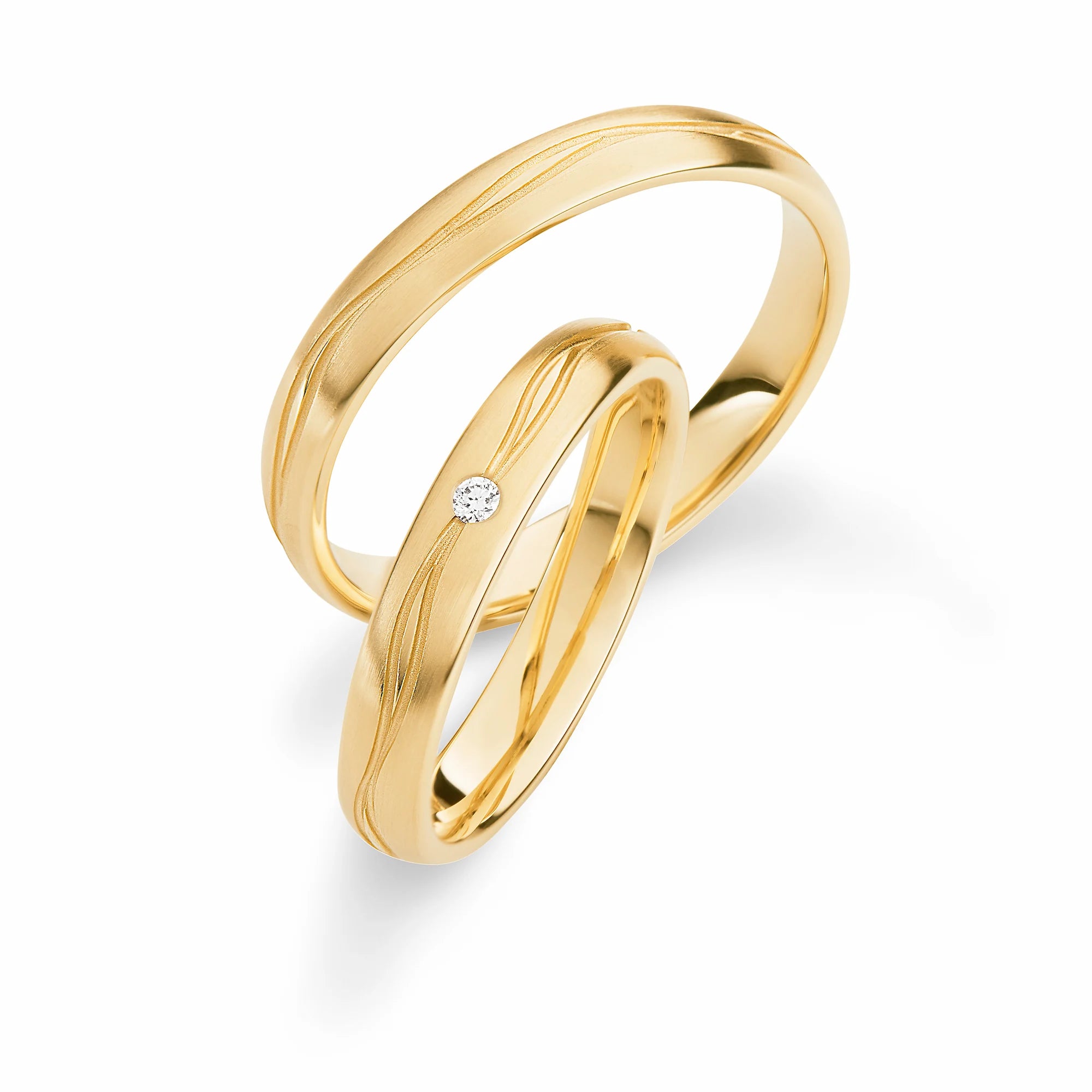 Produktfotografie des gelbgoldenen Max Kemper Trauringpaars mit einer polierten Oberflächenstruktur und zwei wild angeordneten eingravierten Nuten und  einem Diamant im Damenring