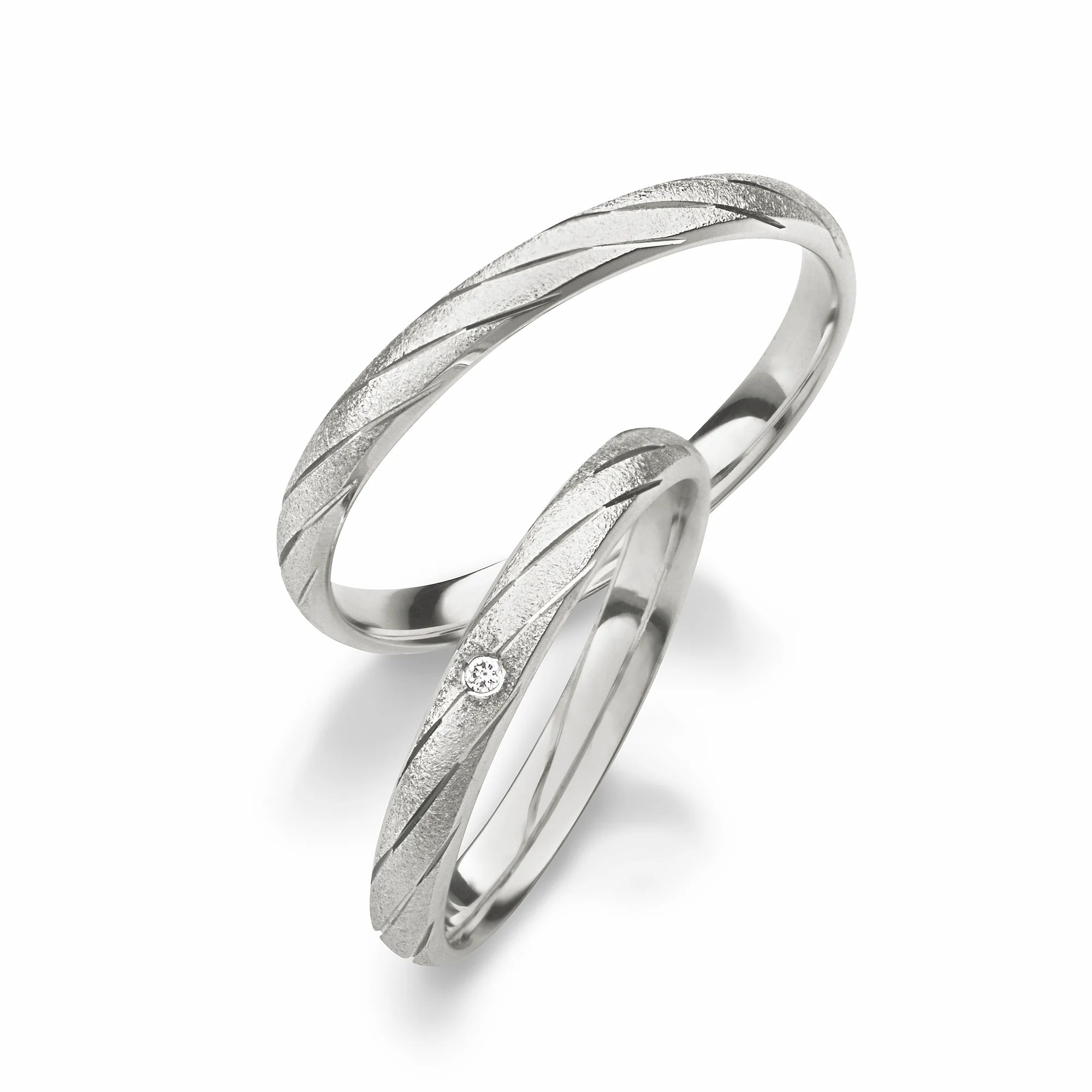 Produktfotografie des weißgoldenen Max Kemper Trauringpaars mit einer einzigartigen Oberflächenstruktur im Frosted-Look mit schräg verlaufenden, eingravierten Nuten und speziell für den Damenring einem eingefassten Diamanten
