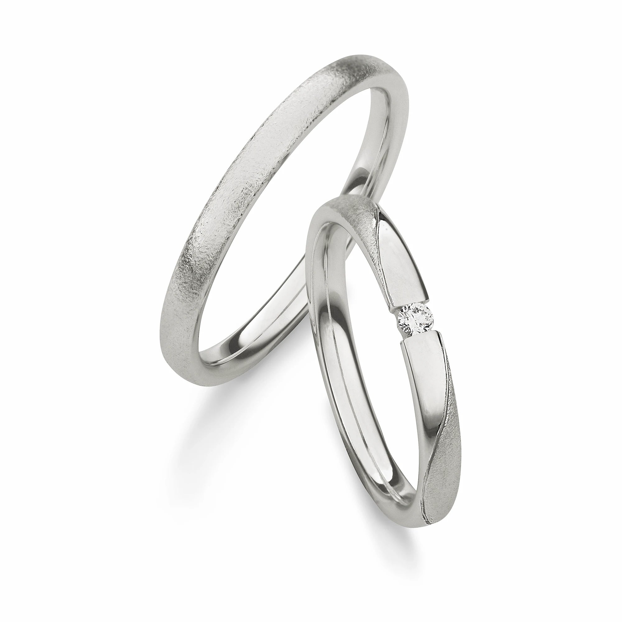 Produktfotografie des weißgoldenen Max Kemper Trauringpaars mit einer leicht angerauten Oberflächenstruktur und speziell für den Damenring eingefassten zentralen Diamanten, welcher von einer glatt polierten Fläche eingerahmt wird