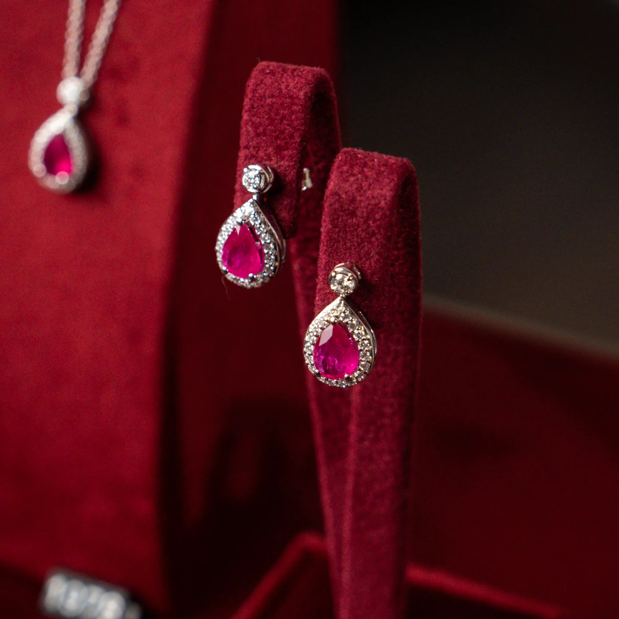 Detailaufnahme der weißgoldenen Ohrstecker von Lunatica Roma mit rotem Rubin im Tropfen-Schliff und Diamant-Besatz, während diese auf einem samtigen, weinroten Juwelier-Ständer präsentiert wird