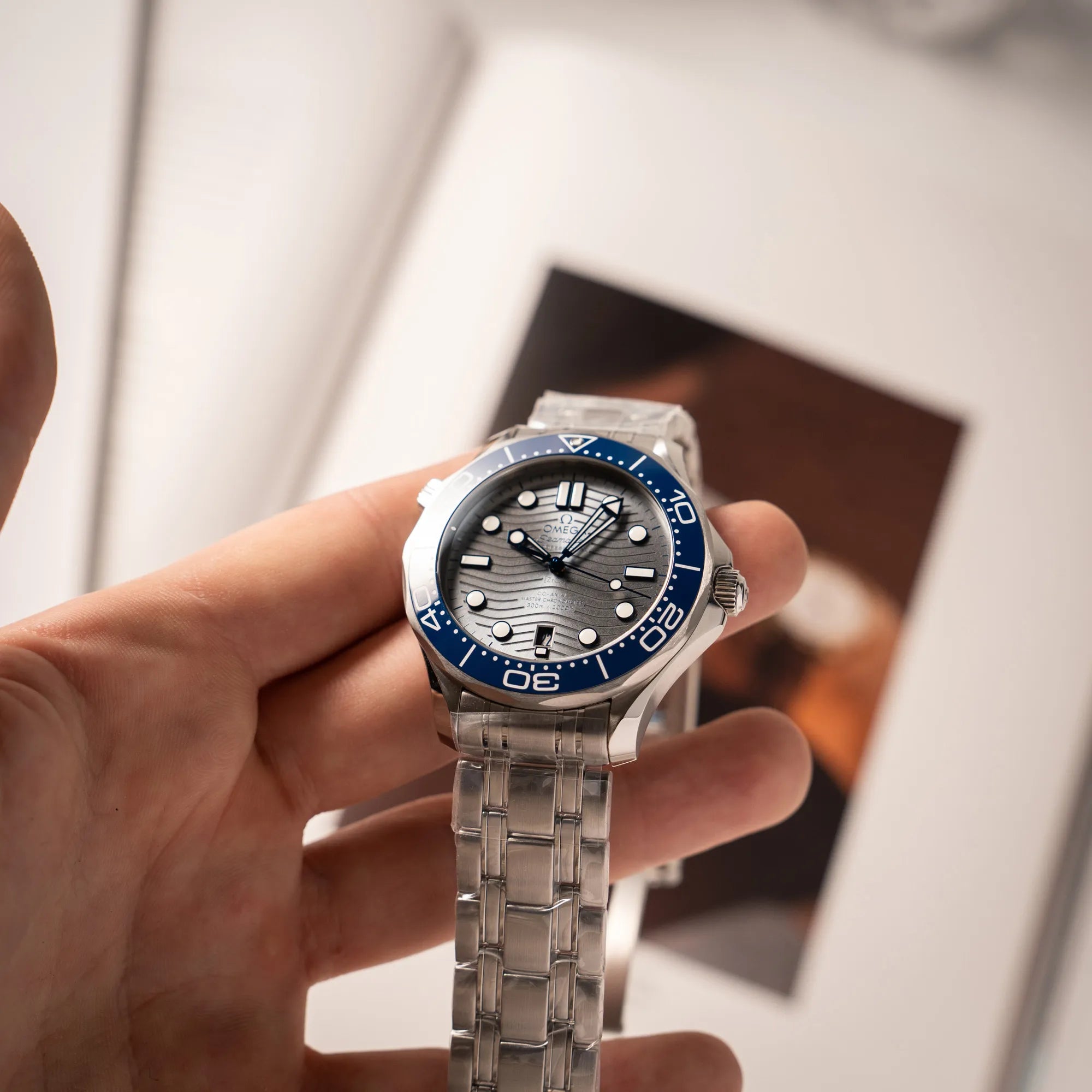Mann hält die Omega Seamaster Diver 300M der Referenz 210.30.42.20.06.001 mit grauem Zifferblatt im Wellen-Design und blauer Lünette am Edelstahlarmband, welches zum Schutz verklebt wurde, in seiner Hand und präsentiert die Uhr in die Kamera