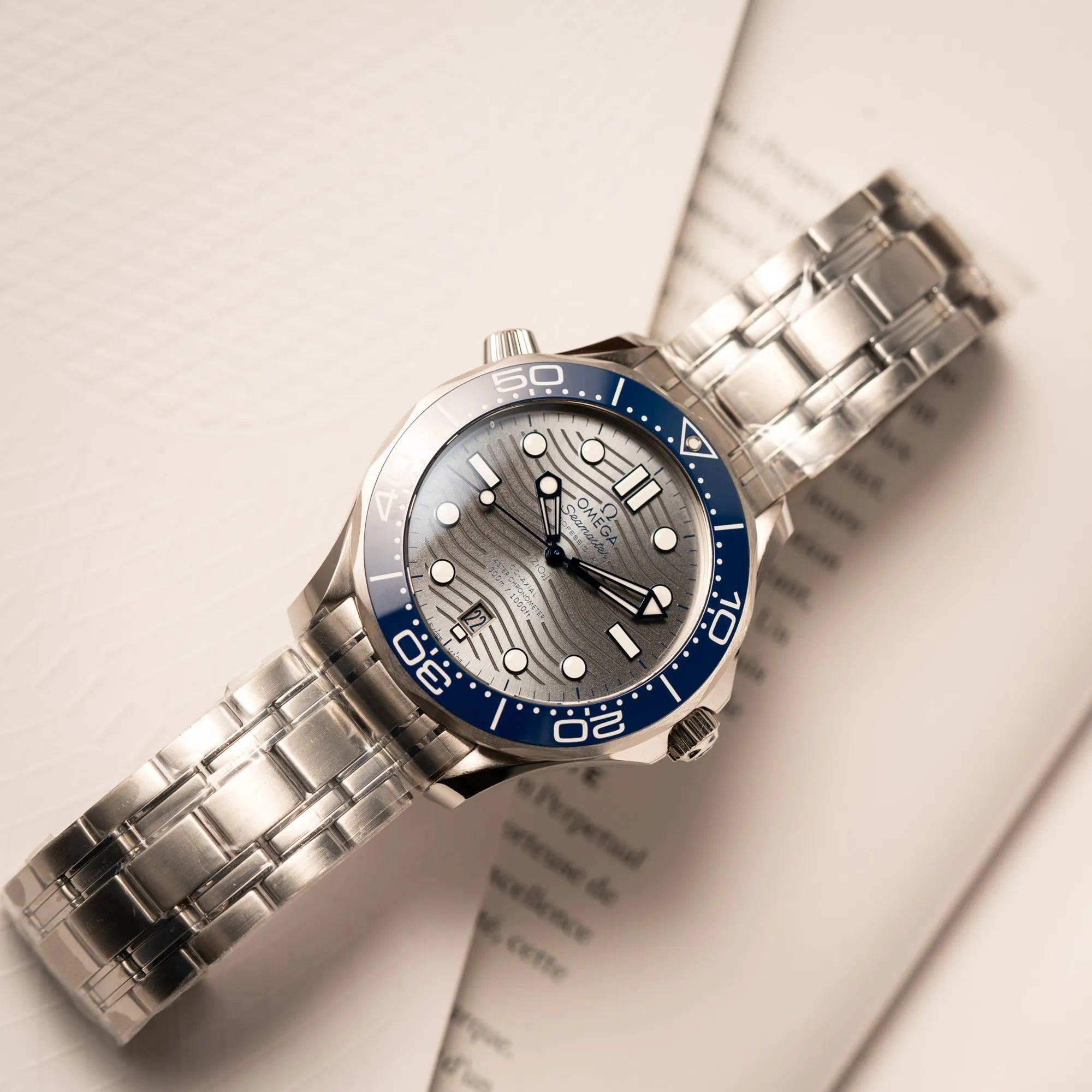 Produktfotografie derOmega Seamaster Diver 300M der Referenz 210.30.42.20.06.001 mit grauem Zifferblatt im Wellen-Design und blauer Lünette am Edelstahlarmband, welches zum Schutz verklebt wurde, zeigt wie die Uhr flach auf einem Buch liegt