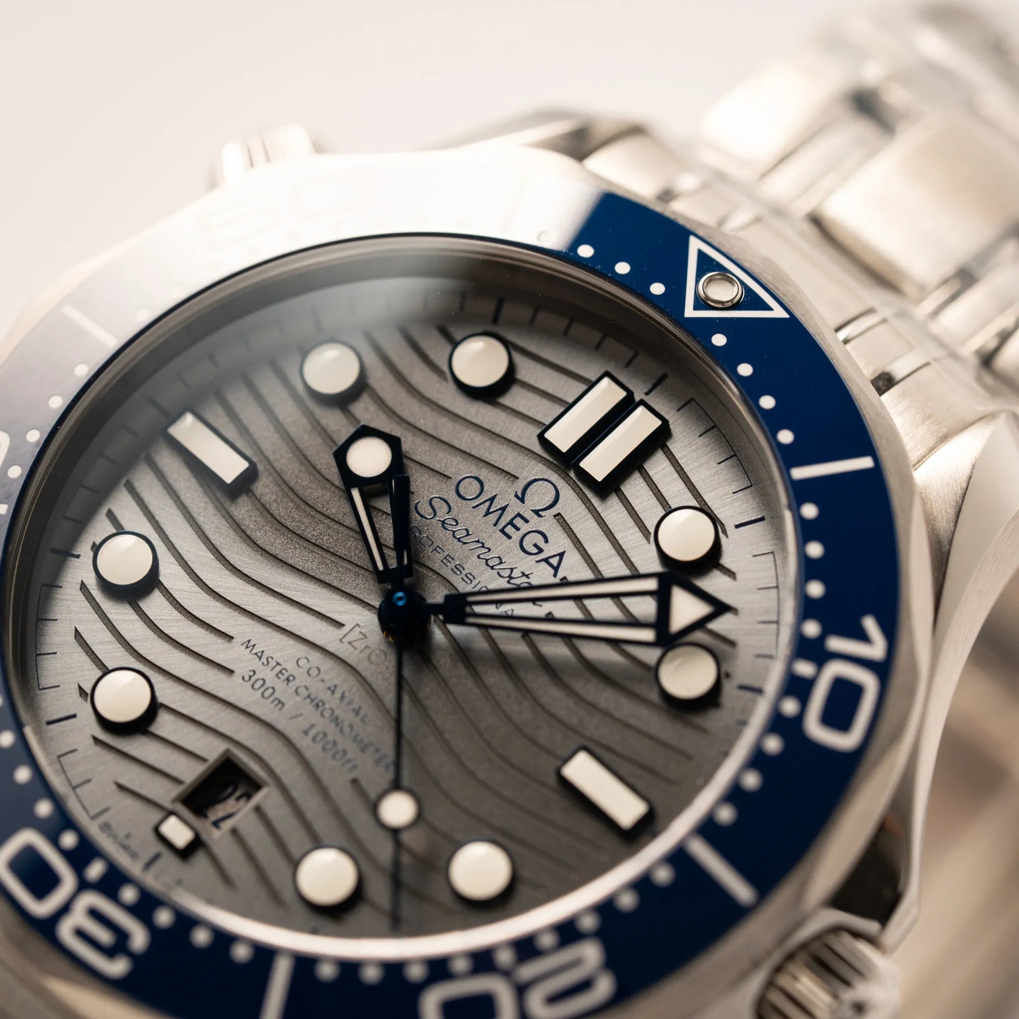 Makroaufnahme des Uhrenkopfes der Omega Seamaster Diver 300M der Referenz 210.30.42.20.06.001 mit grauem Zifferblatt im Wellen-Design und blauer Lünette am Edelstahlarmband, welches zum Schutz verklebt wurde