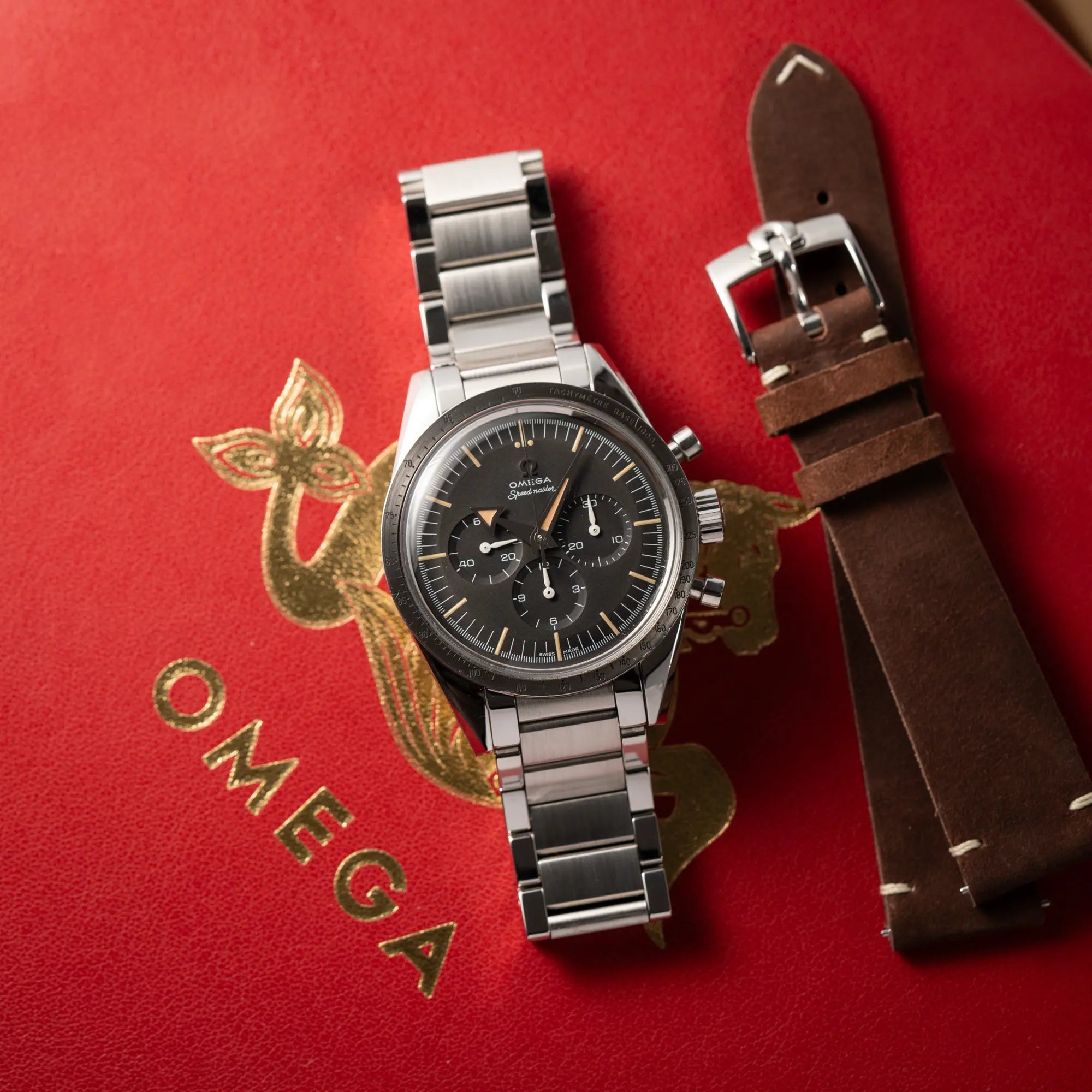 Produktfotografie der Omega Speedmaster 1957 Trilogy Limited Edition mit einem anthrazitfarbenen Zifferblatt mit Vintage-Look und dem original Omega-Stahlband im Ed-White-Design zusammen mit dem zusätzlichen braunem Omega-Lederband, während die Uhr auf der roten Omega-Box liegt