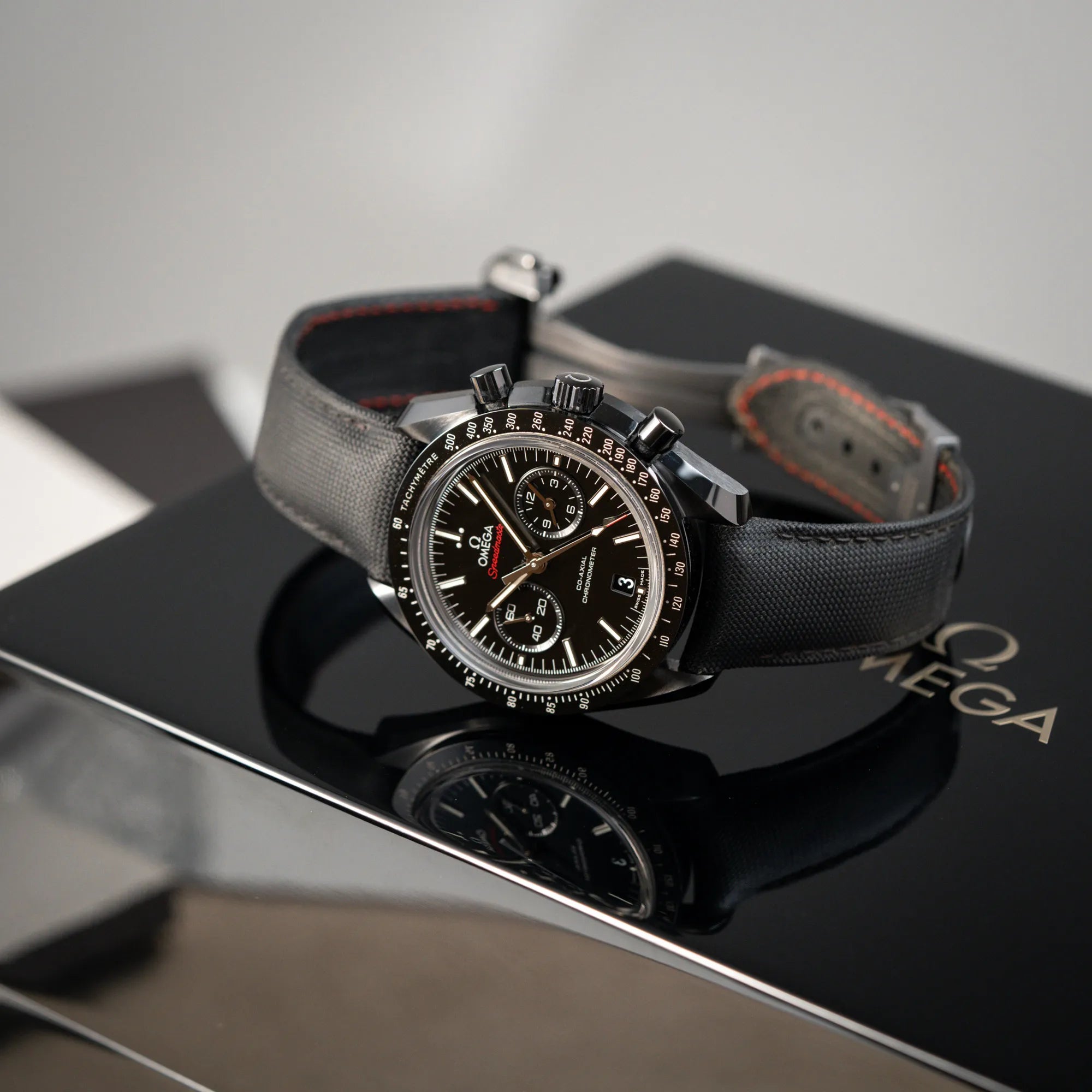 Produktfotografie des auf der Uhrenbox liegenden Omega Speedmaster "Darkside of the Moon" Chronograph mit schwarzem Zifferblatt und aus schwarzer Keramik gefertigtem Gehäuse