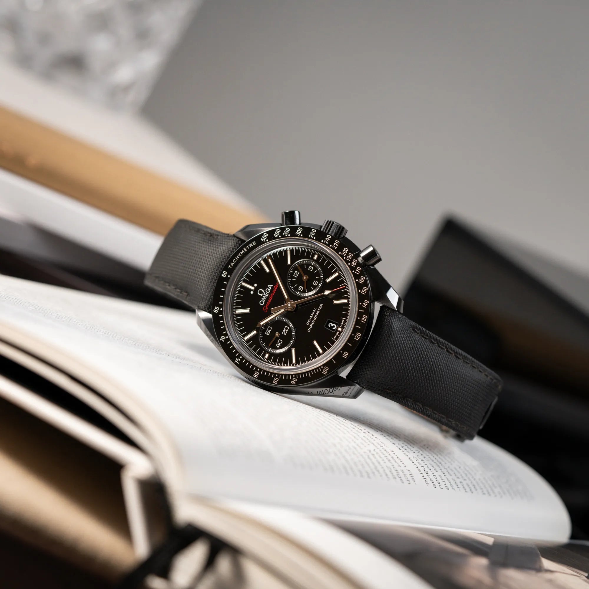 Produktfotografie des Omega Speedmaster "Darkside of the Moon" Chronograph mit schwarzem Zifferblatt und aus schwarzer Keramik gefertigtem Gehäuse, während die Uhr auf einem Buch liegt