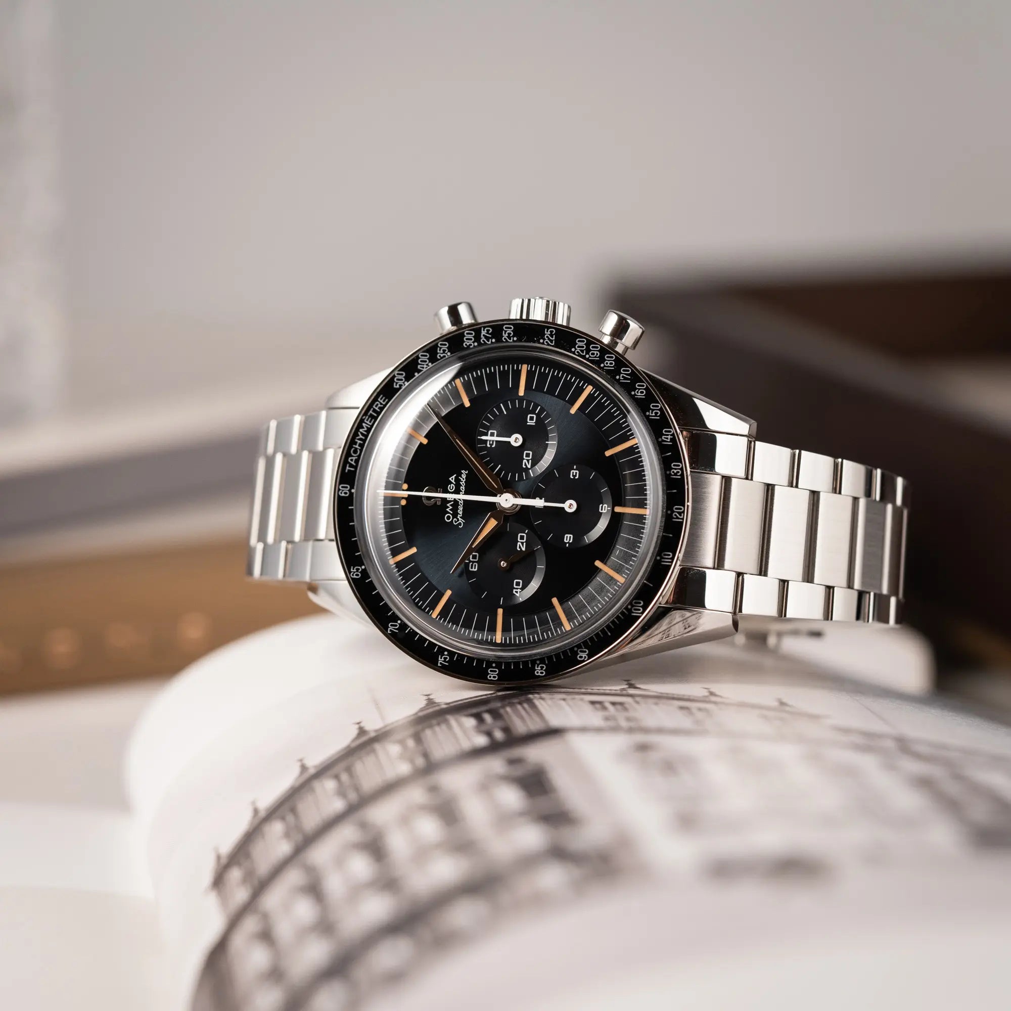 Produktfotografie der seitlich auf dem Uhrengehäuse liegenden Omega Speedmaster FOIS der Referenz 310.30.40.50.06.001 mit dem dunkelblauen Zifferblatt mit dunkel-beigen Indizes im Vintage-Look und einem vom Standard-Modell abgeänderten Stahl-Armband, was beides an das klassische Speedy-Modell  erinnert